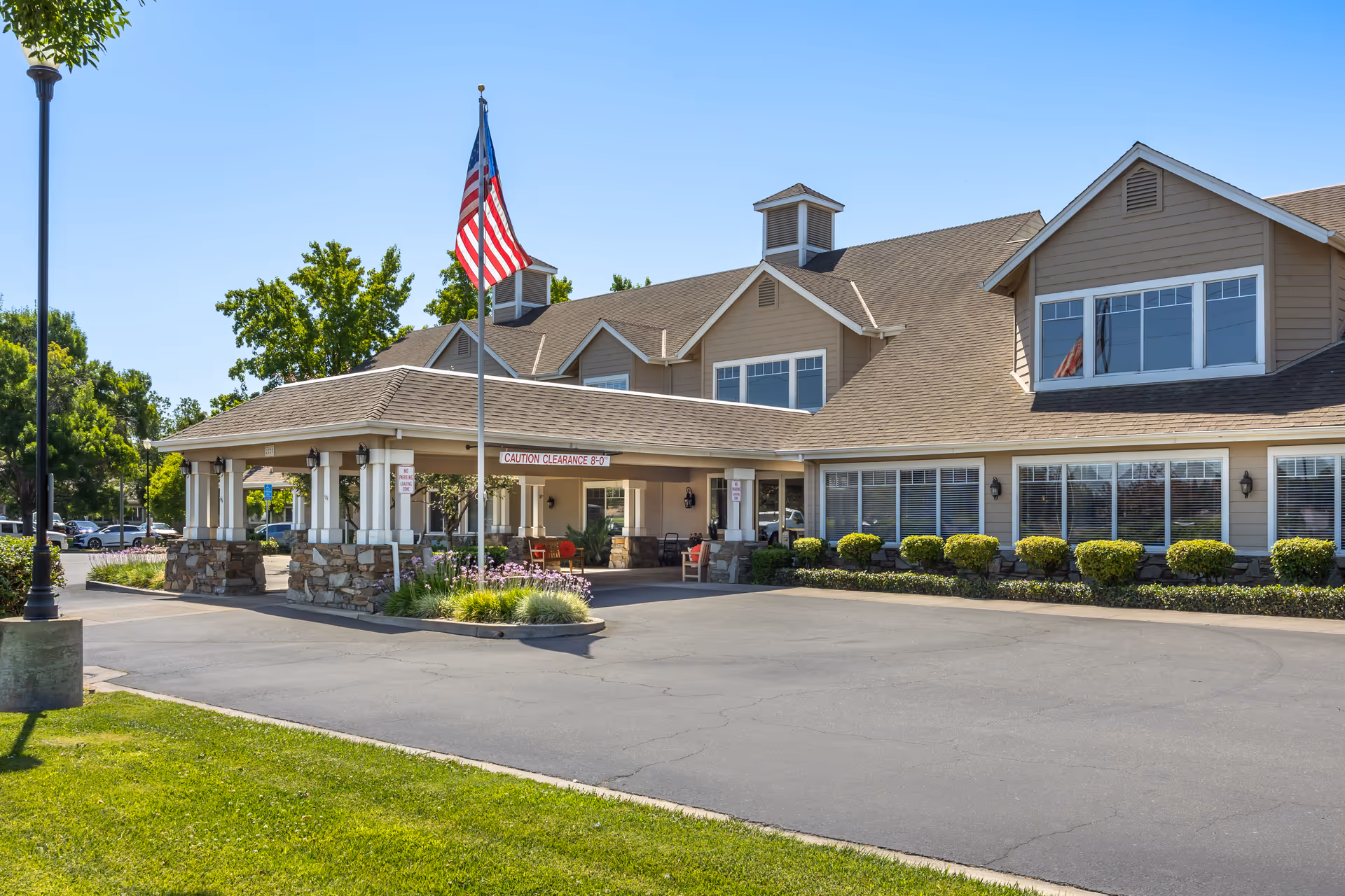 Exterior view of Brookdale Lodi senior living facility showing a large building with beige siding and multiple windows. There is a covered entrance with stone pillars, an American flag on a flagpole, and neatly trimmed bushes along the front. The sky is clear and blue.