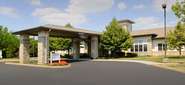 Front entrance of the Sanctuary At Tuttle Crossing with a covered porte-cochère, stone columns, driveway, and landscaped trees and shrubs.