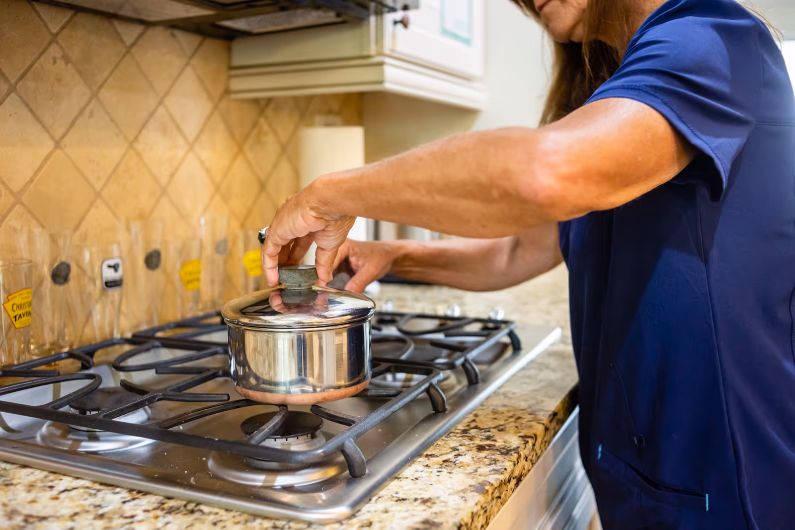 A caregiver in blue scrubs cooks on a stove with a pot.