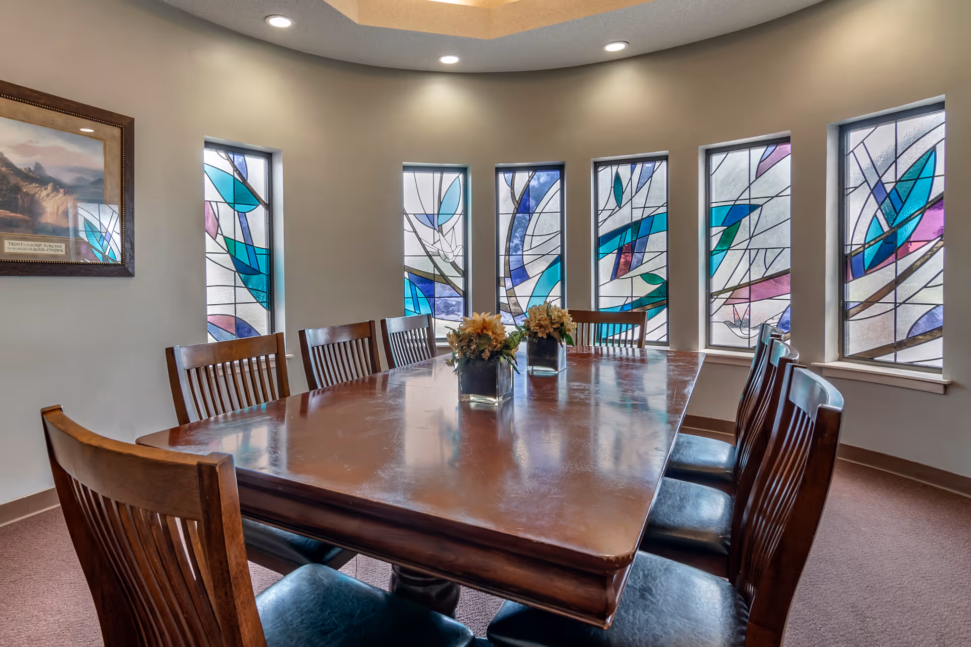 A dining room with a large polished wooden table surrounded by wooden chairs with black cushions. The room features six tall stained glass windows with colorful abstract designs, and two small flower arrangements in square glass vases are placed on the table. The walls are painted beige and the ceiling has recessed lighting.