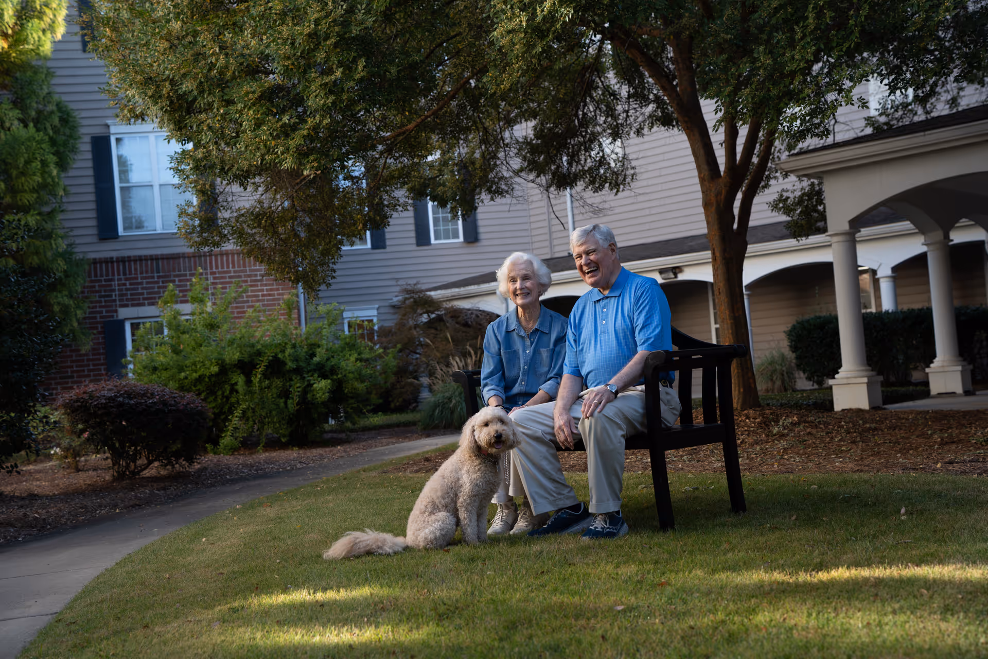 An elderly couple sitting on a bench outside near a tree with a light brown dog sitting on the grass in front of them. They are smiling and appear to be enjoying a pleasant day at the facility named Spring Harbor at Green Island. The background shows part of the building with windows, bushes, and a covered walkway supported by columns.