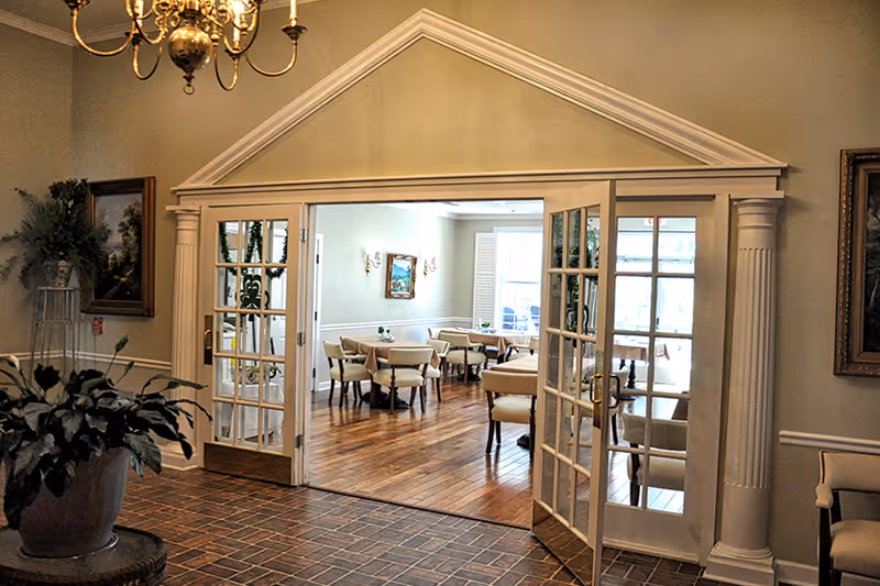 View through open double glass doors into a dining room with several tables and chairs arranged neatly. The room has wooden flooring, light-colored walls with paintings, and wall sconces. In the foreground, there is a tiled floor area with a large potted plant and a chandelier hanging from the ceiling.
