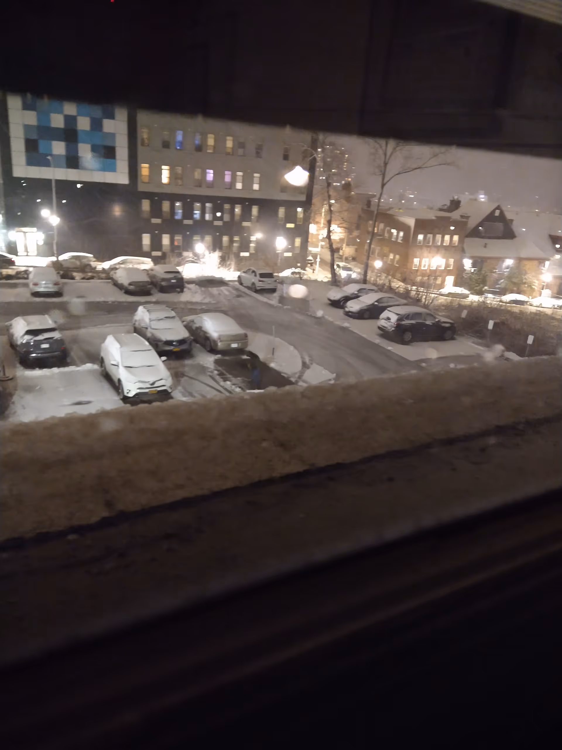 Snowy night exterior view of a building complex with parked cars, seen from a window.