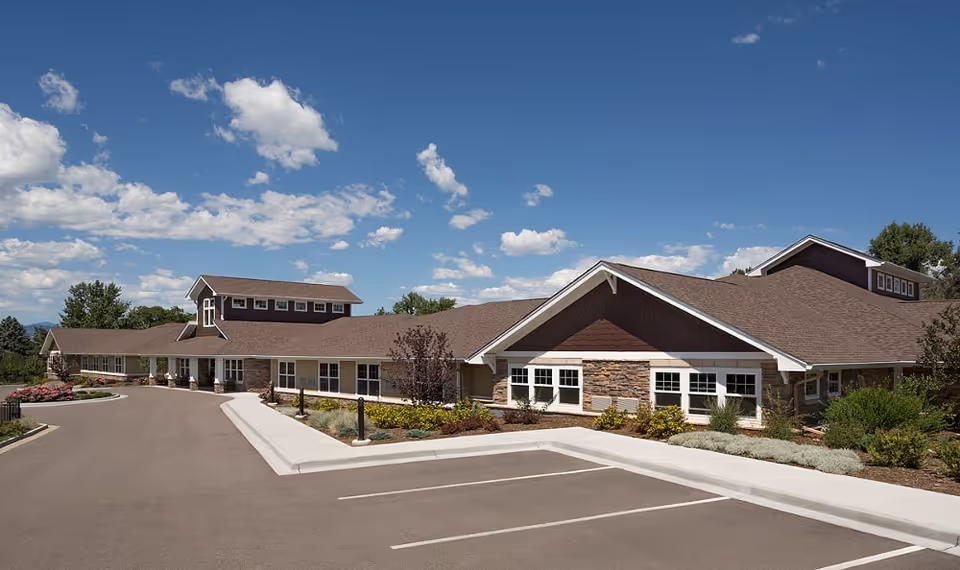 Exterior view of a single-story senior living facility building with a brown roof, beige walls, and stone accents. The building is surrounded by landscaped greenery and a clear blue sky with scattered clouds is visible above.