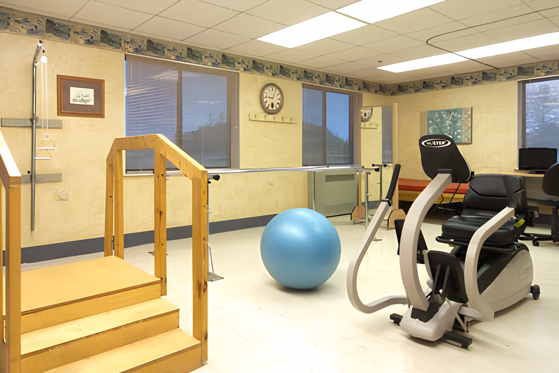 Rehabilitation exercise room with wooden practice stairs, parallel bars, a blue exercise ball, and a recumbent exercise machine.