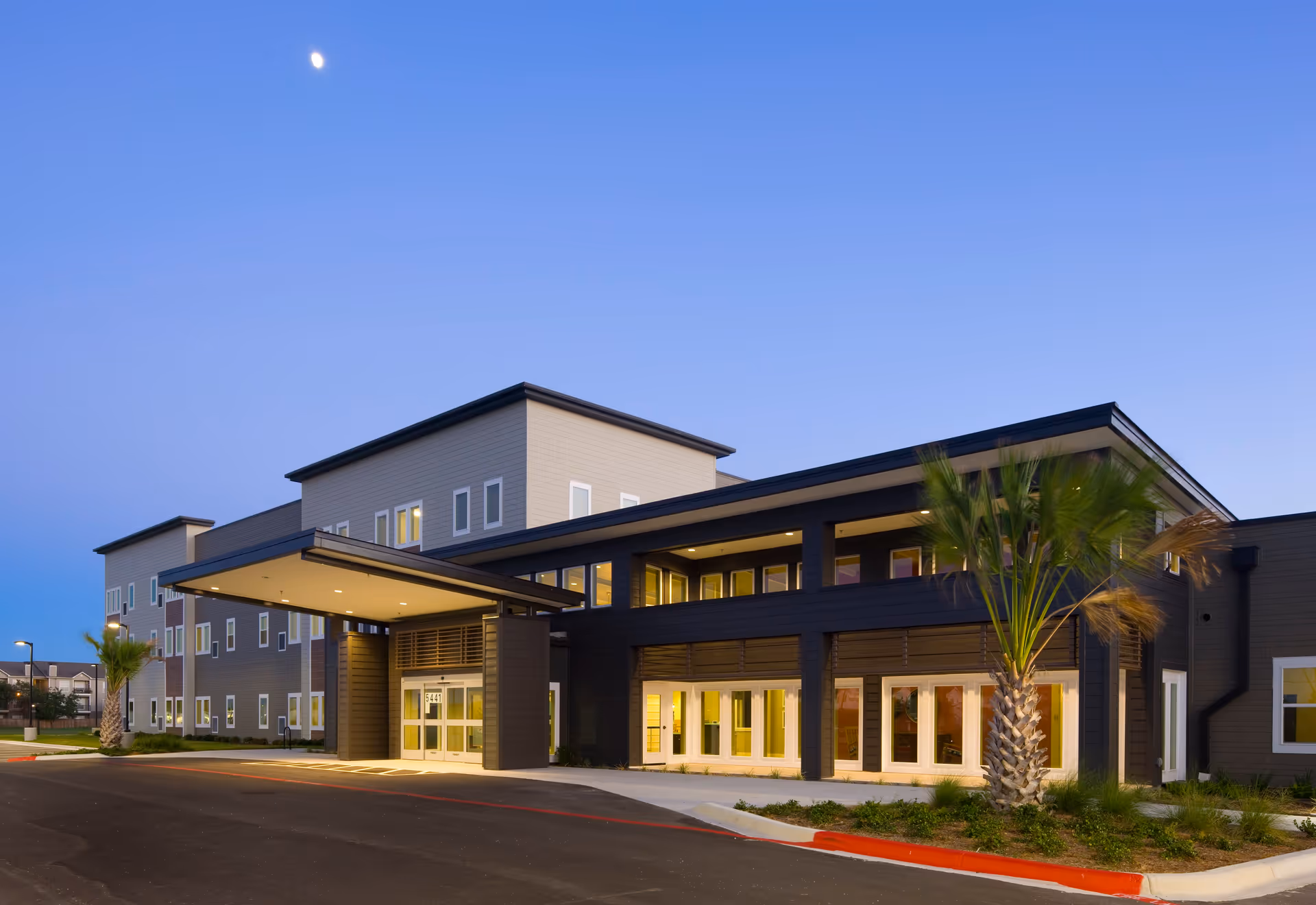 Exterior view of a modern senior living facility building at dusk with illuminated windows, a covered entrance, palm trees, and a clear sky.
