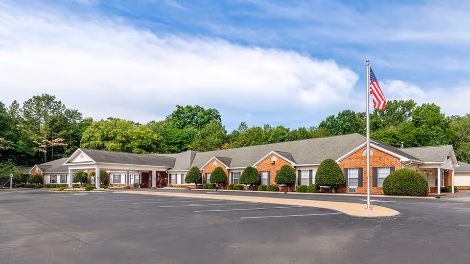 Single-story brick senior living facility building with a flagpole and empty parking lot in front, surrounded by trees.