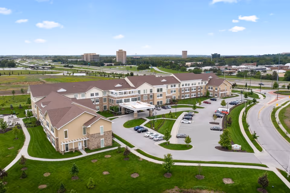 Aerial view of a large senior living facility building with beige and stone exterior, surrounded by green lawns, trees, and a parking lot with several cars. The building has multiple wings and a covered entrance. Roads and highways are visible in the background under a partly cloudy blue sky.