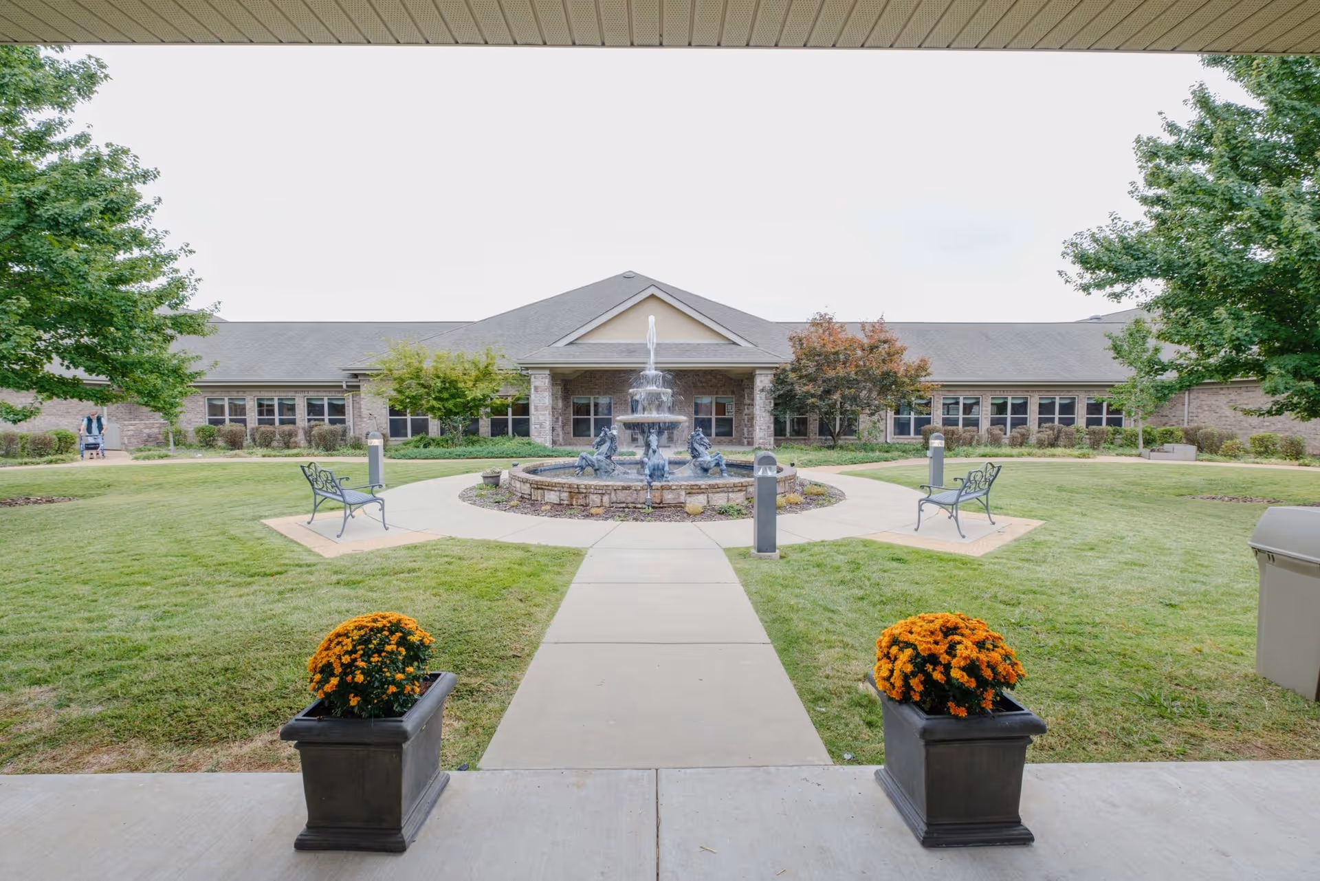 Covered entryway looking out onto a landscaped courtyard with a central fountain, benches, and the facility building beyond.