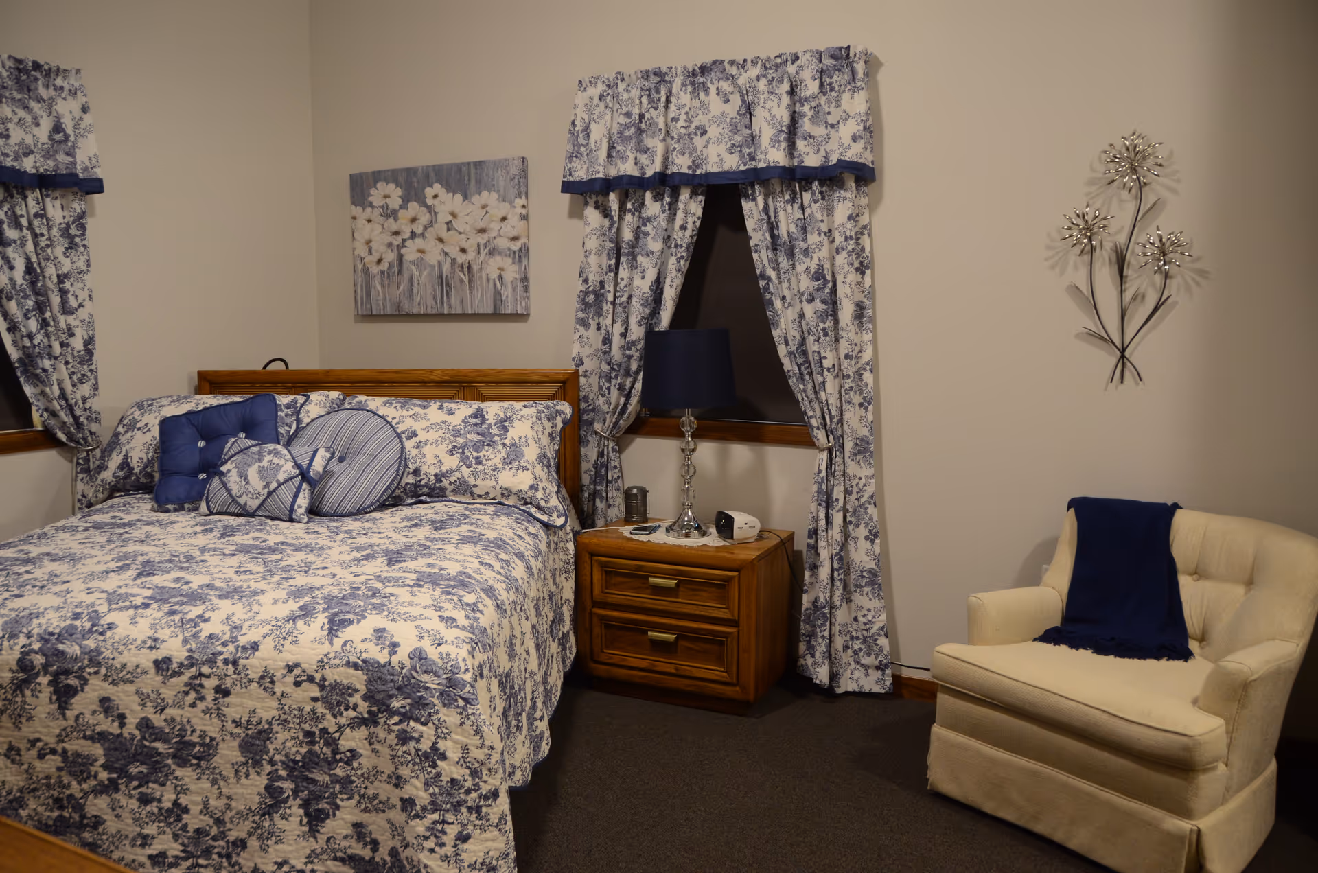 A blue floral bedroom with a bed, curtains, nightstand, lamp, and beige armchair.