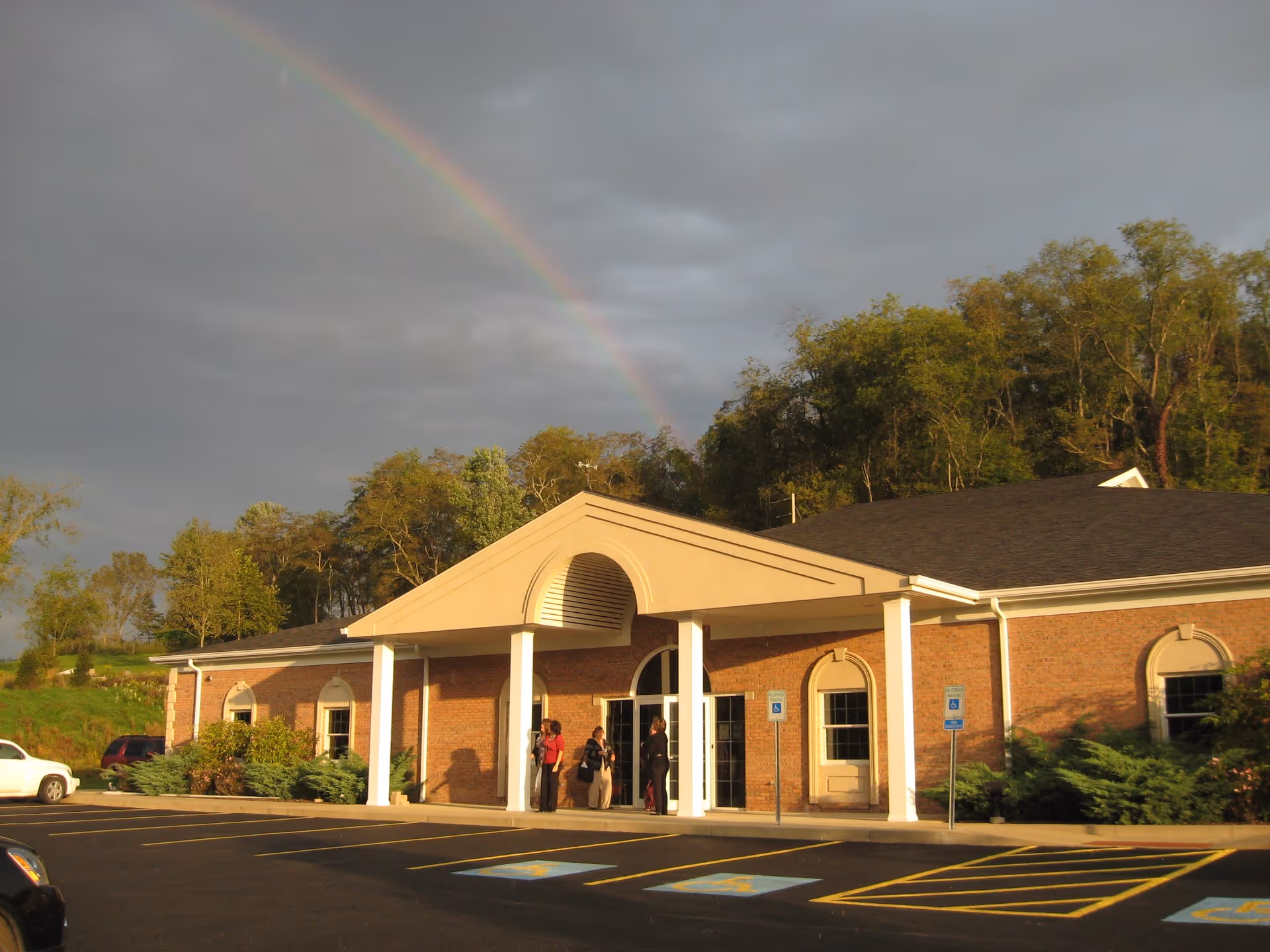Brick Valley Hospice building with a front entrance, rainbow, and people outside.
