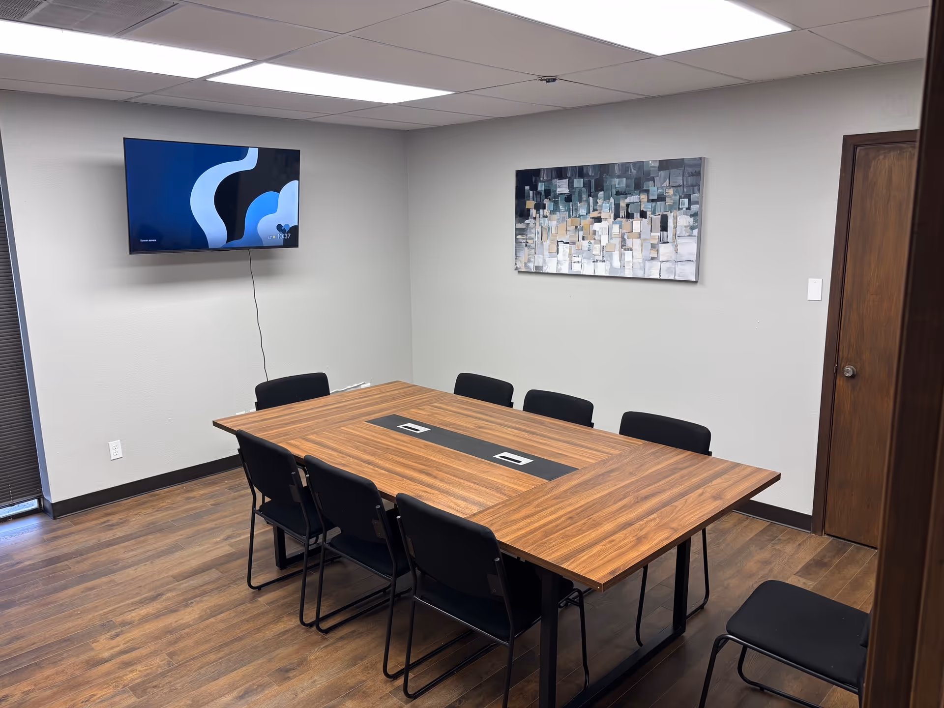 A conference room with a wooden table, black chairs, wall-mounted TV, and abstract art.