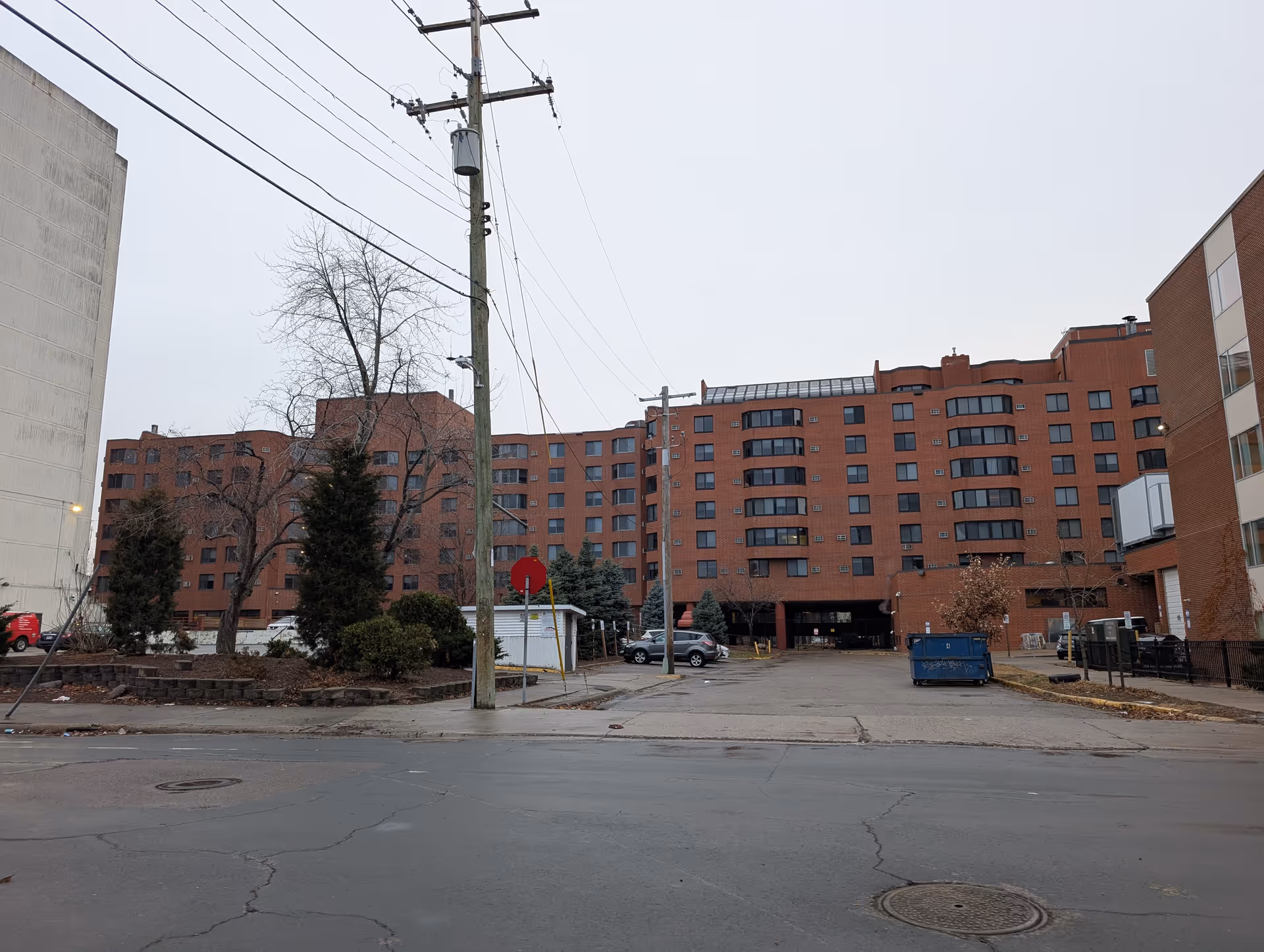 Exterior view of a large multi-story brick building with multiple windows, a parking area in front with a few cars and a blue dumpster, and some leafless trees and shrubs around the property under an overcast sky.