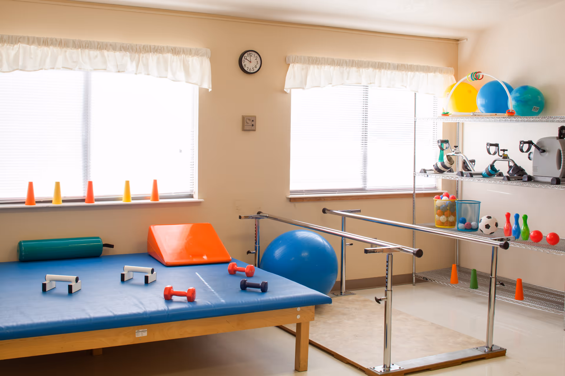 Indoor physical therapy room with a blue padded table, parallel bars, a large blue exercise ball, and shelves stocked with therapy equipment.