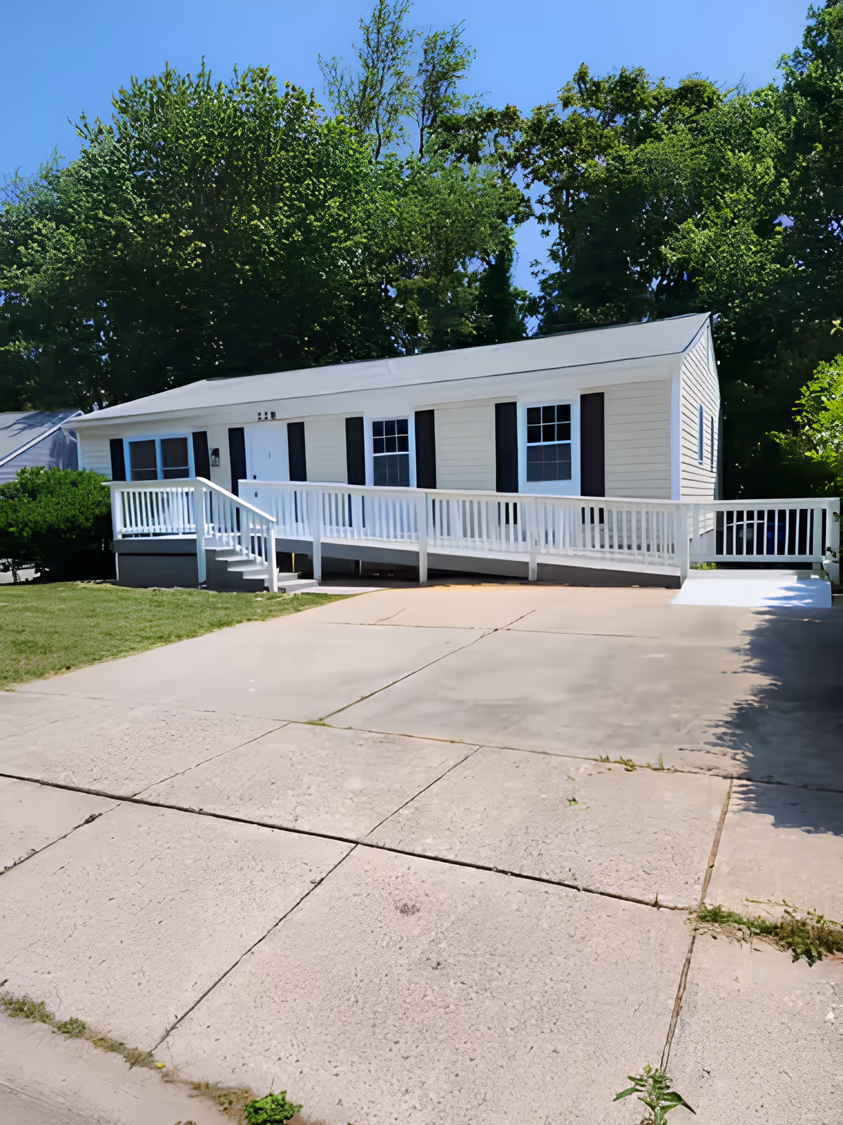 Single-story manufactured home with a white wheelchair ramp and driveway, set against tall trees and a clear sky.