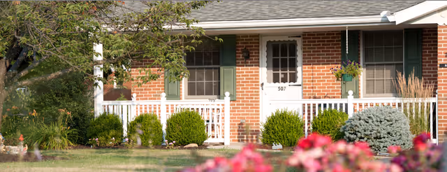 Front exterior of a brick single-story residence with a white porch railing, green shutters, house number 507, and landscaped shrubs and flowers.