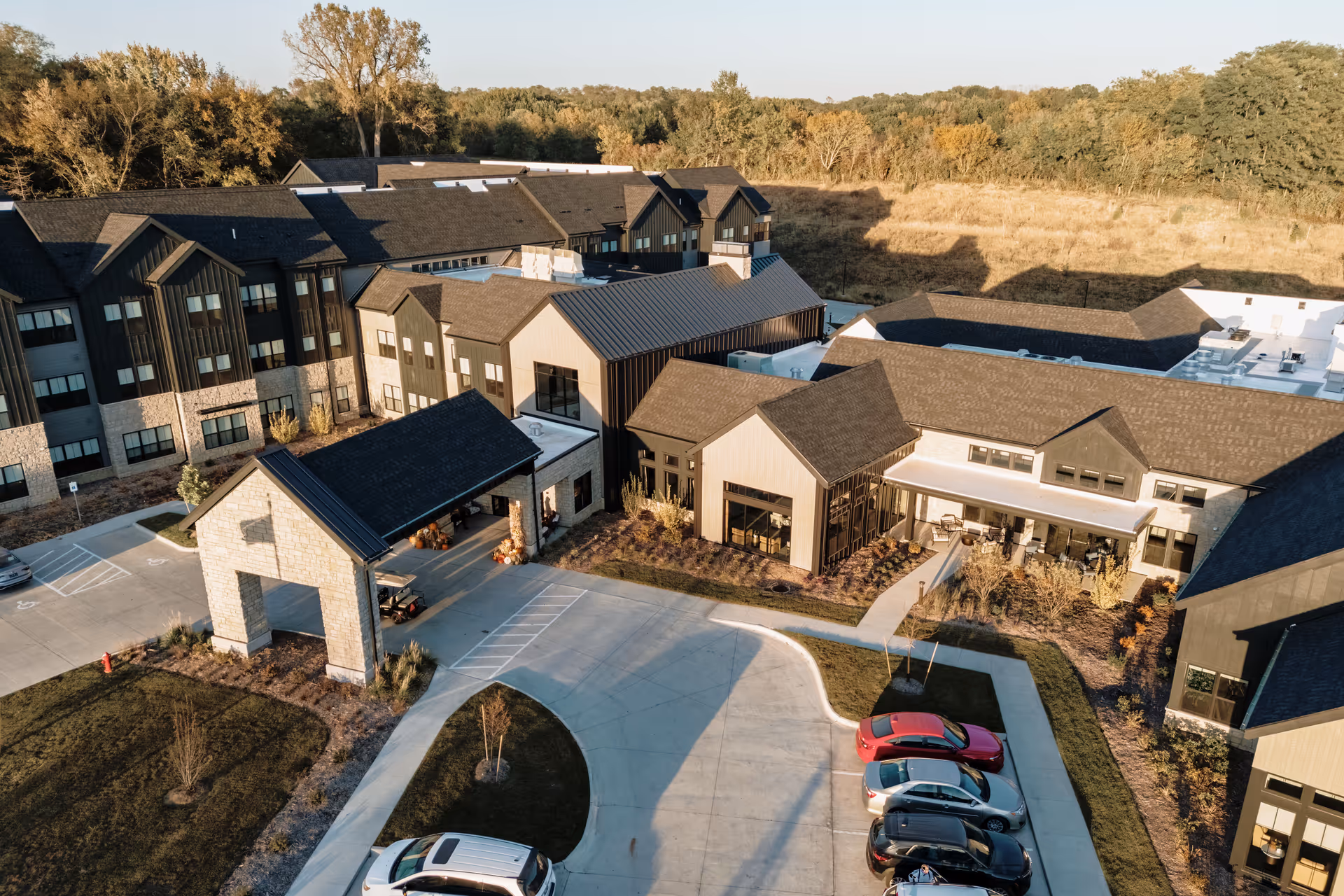 Aerial view of a senior living facility with connected modern buildings, a covered entrance porte-cochere, parked cars, and surrounding trees.