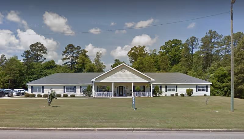 Single-story white facility with a front porch, lawn, and trees behind.