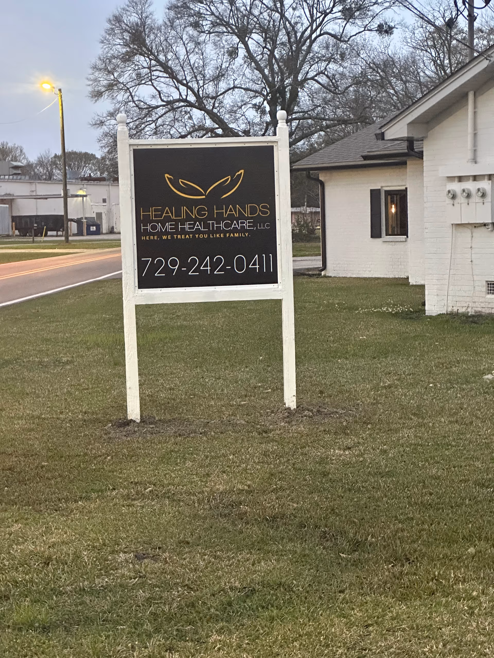 Outdoor sign for Healing Hands Home Healthcare LLC beside a white brick building and trees nearby.