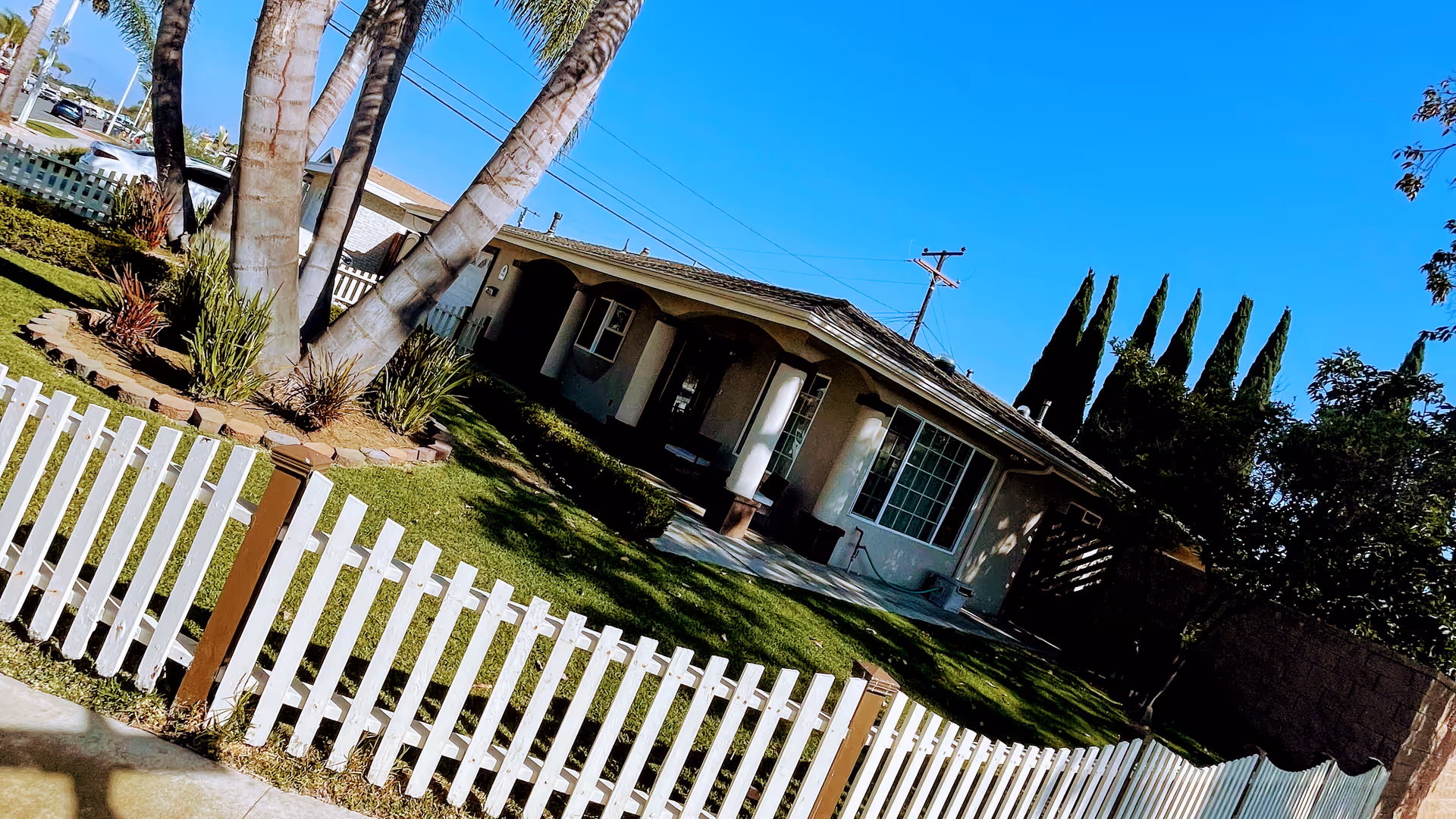 Single-story senior living facility with white picket fence, green lawn, and palm trees in front.