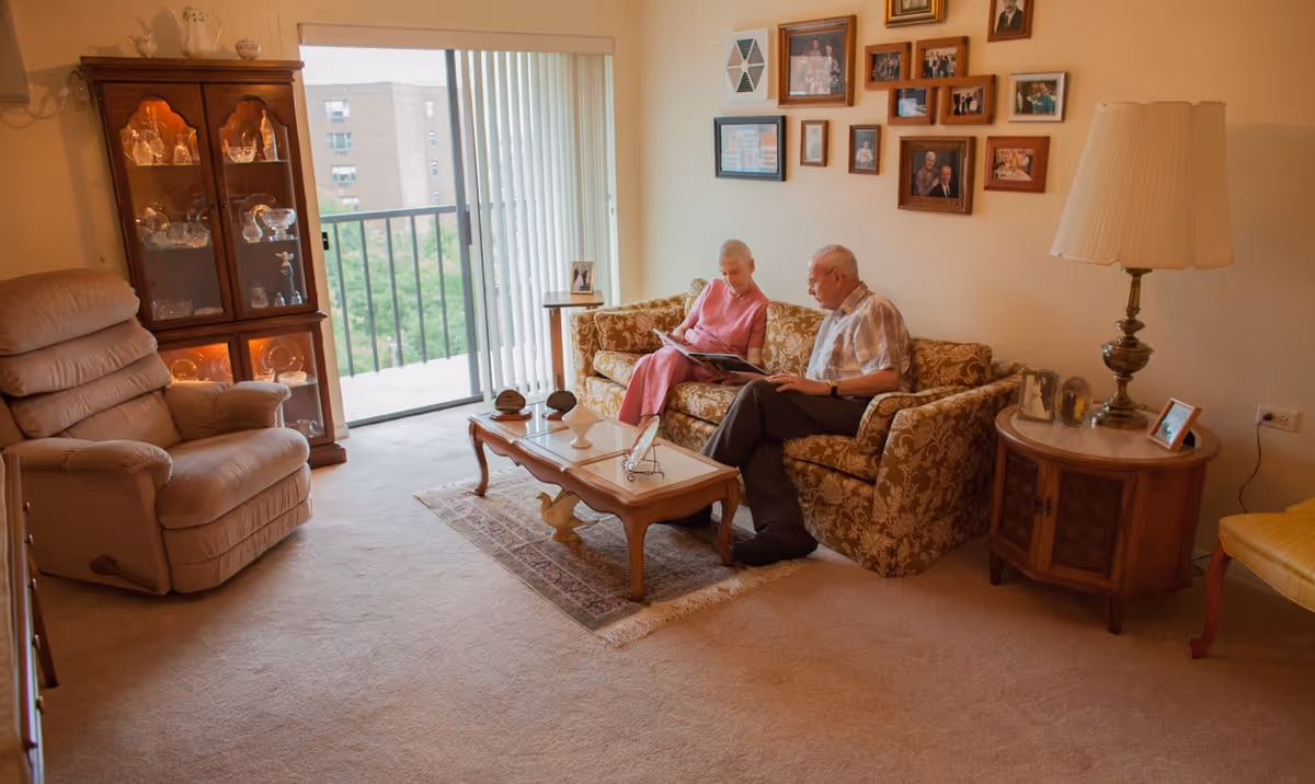 Living room with two people sitting on a patterned sofa reading over a coffee table, with a recliner, display cabinet, side table and sliding glass door visible.