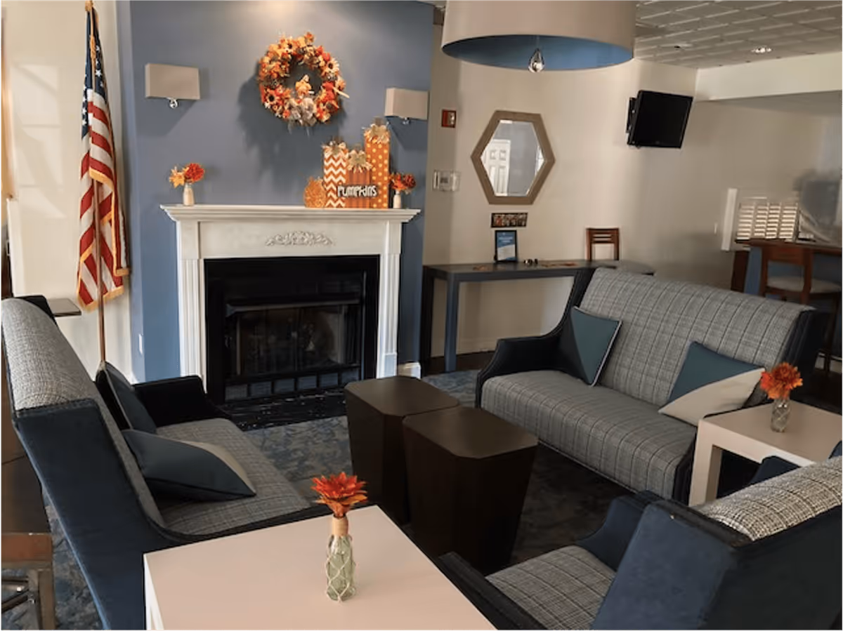 A cozy living room area with a fireplace decorated with a fall-themed wreath and pumpkins. The room has a blue accent wall, an American flag, a hexagonal mirror, a wall-mounted TV, and seating including two armchairs and a sofa with plaid upholstery and decorative pillows. Small tables with flower vases are placed near the seating.