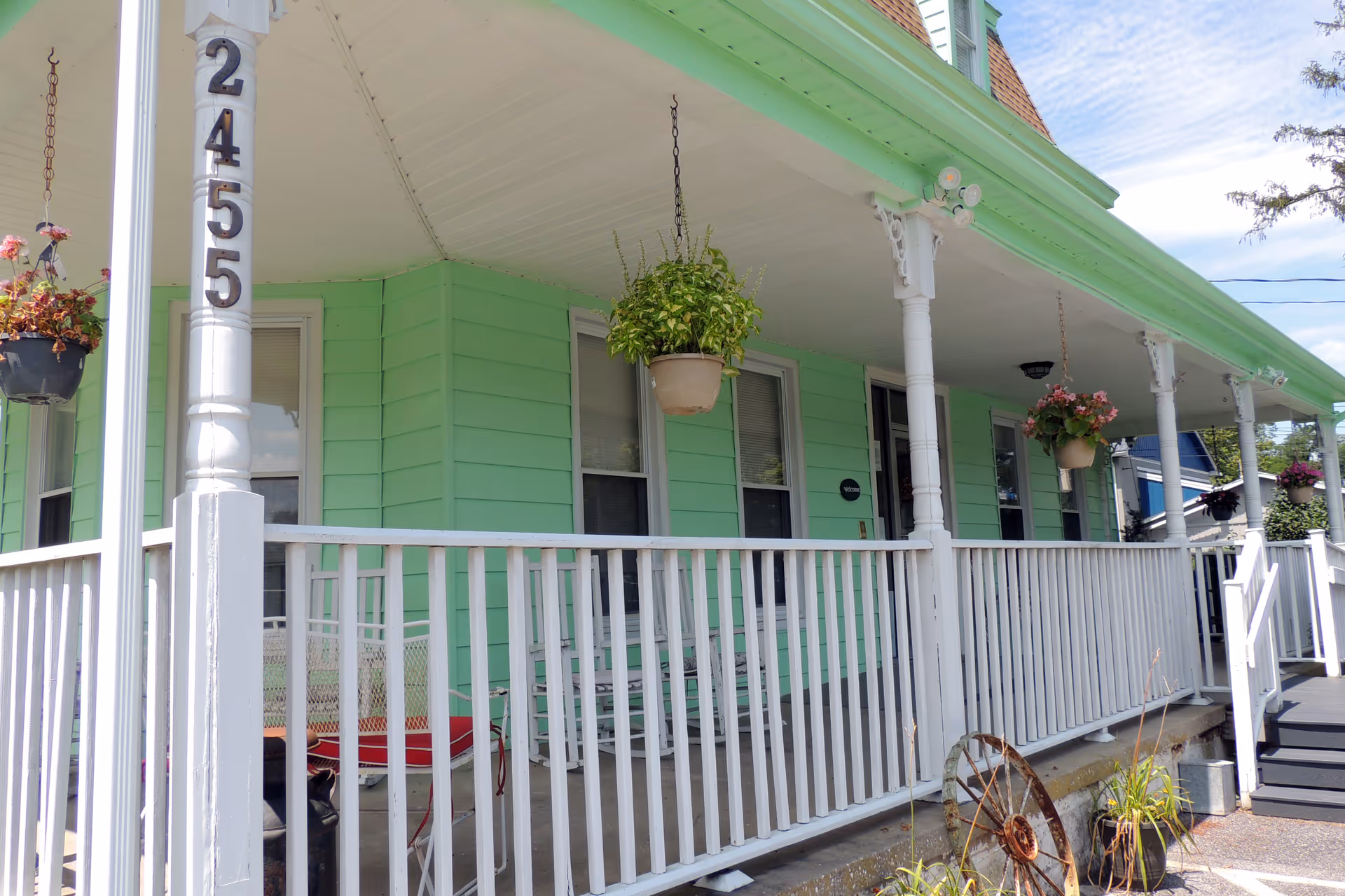 Front porch of a light green house with white railings and columns, hanging flower pots, and the house number 2455 displayed on a column.