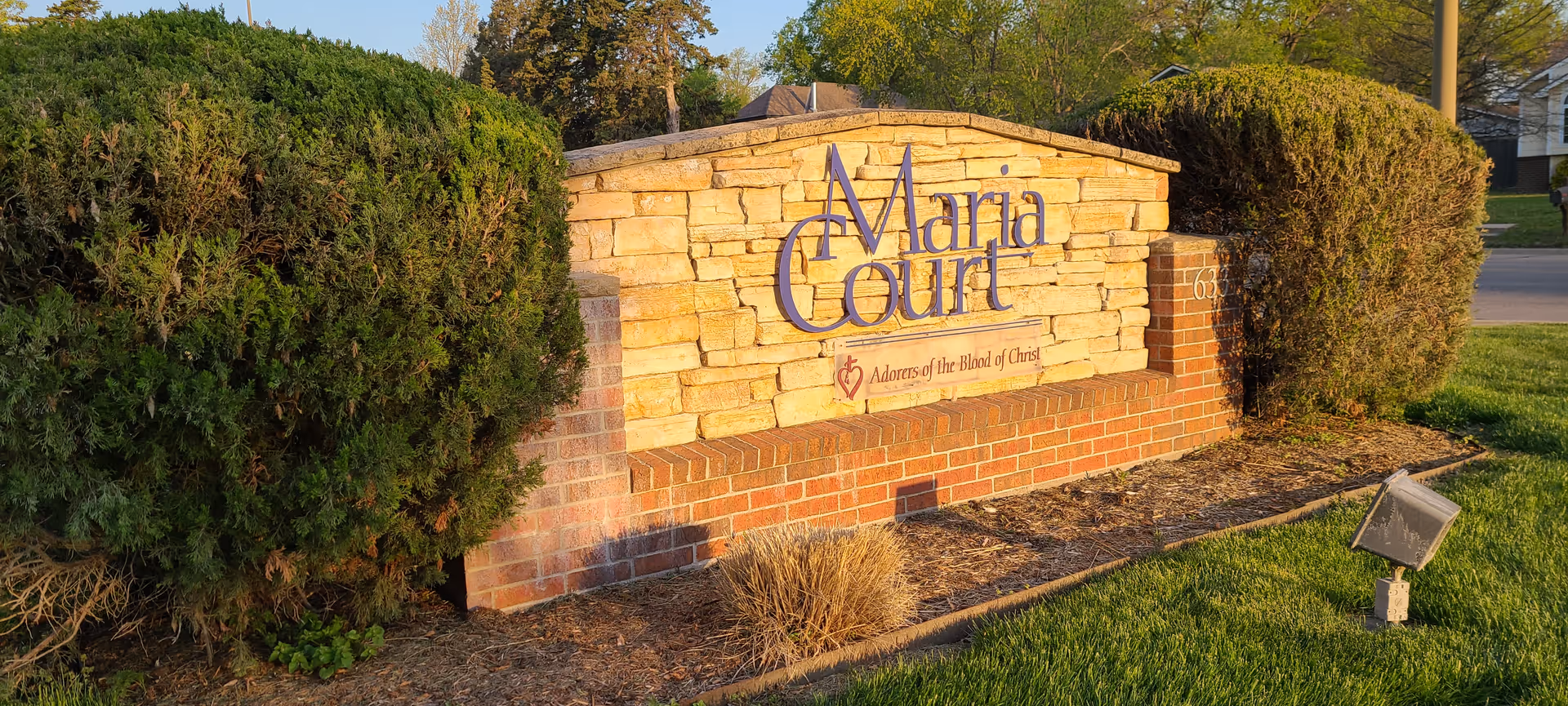 Outdoor stone and brick sign for Maria Court with the phrase 'Adorers of the Blood of Christ' beneath the name, surrounded by trimmed bushes and grass in a residential area.