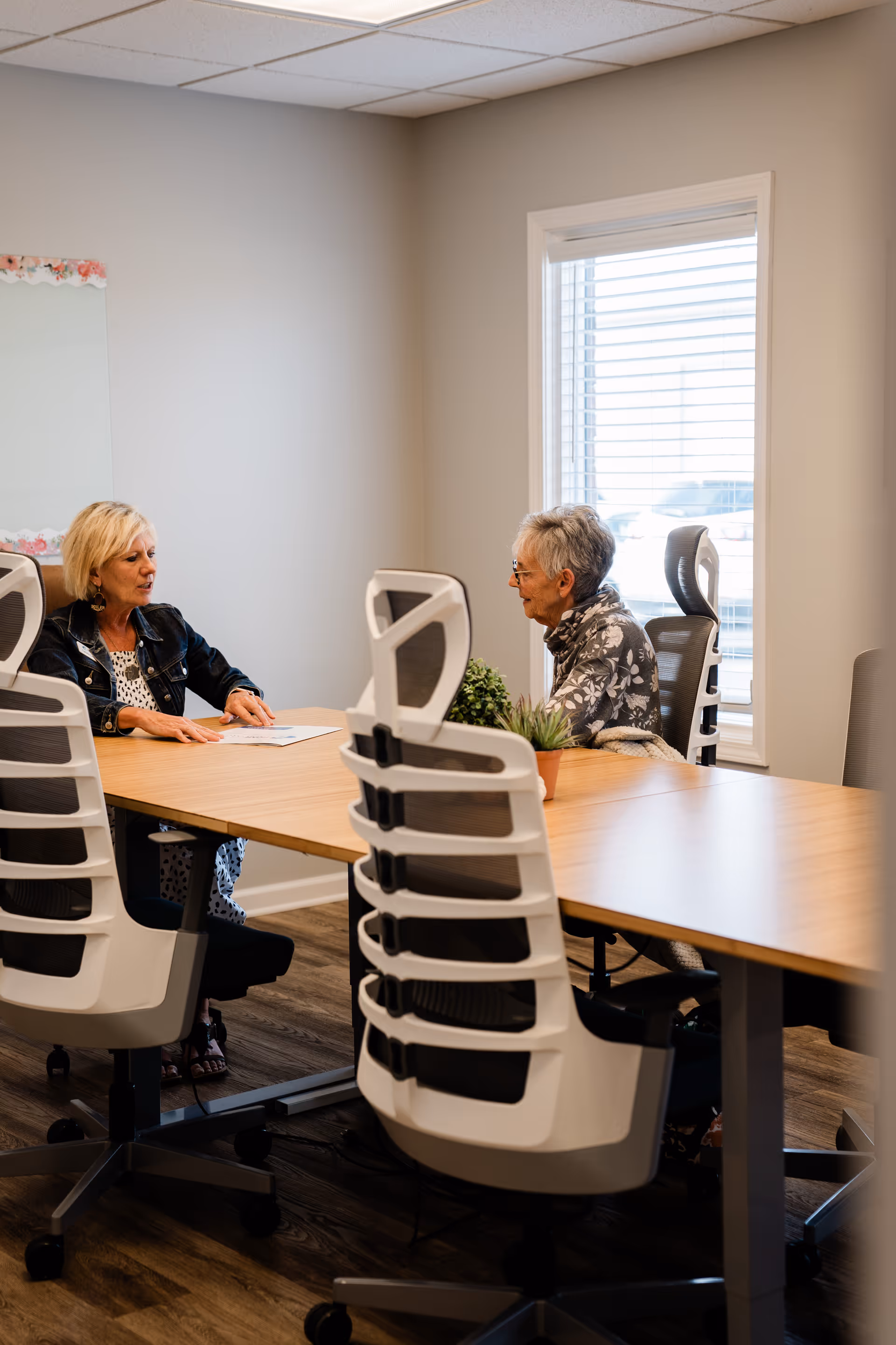 Two elderly women sit at a long conference table in a modern office-like room.