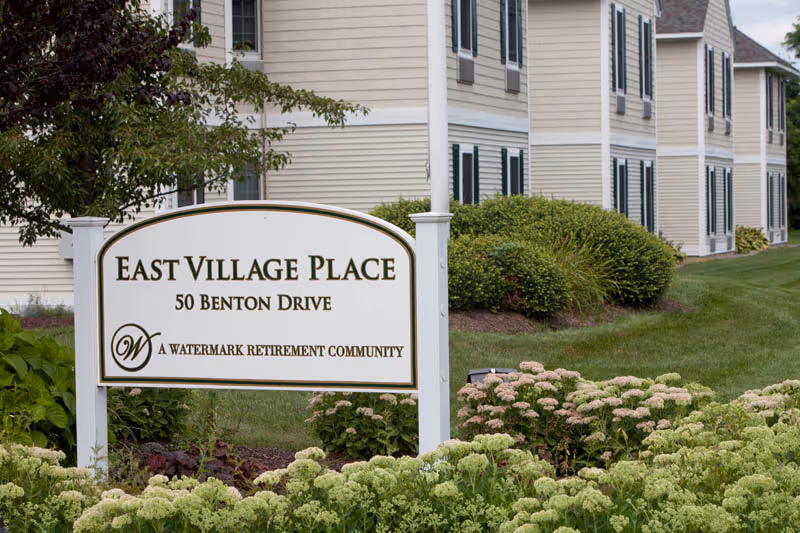 A landscaped entrance sign reading "East Village Place, 50 Benton Drive" in front of a beige multi-unit building.
