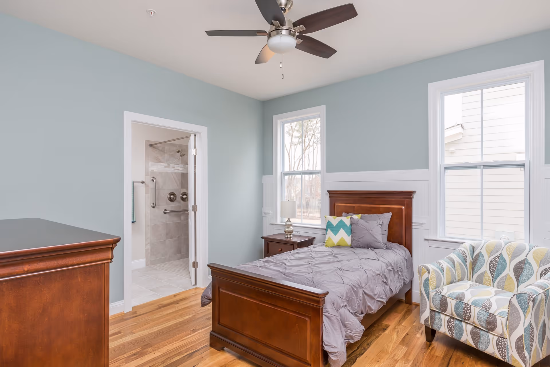 A bedroom with light blue walls and hardwood floors featuring a wooden bed with gray bedding and colorful pillows, a matching wooden nightstand with a lamp, a patterned armchair, and two large windows letting in natural light. An open door reveals a tiled bathroom with a walk-in shower and grab bars.