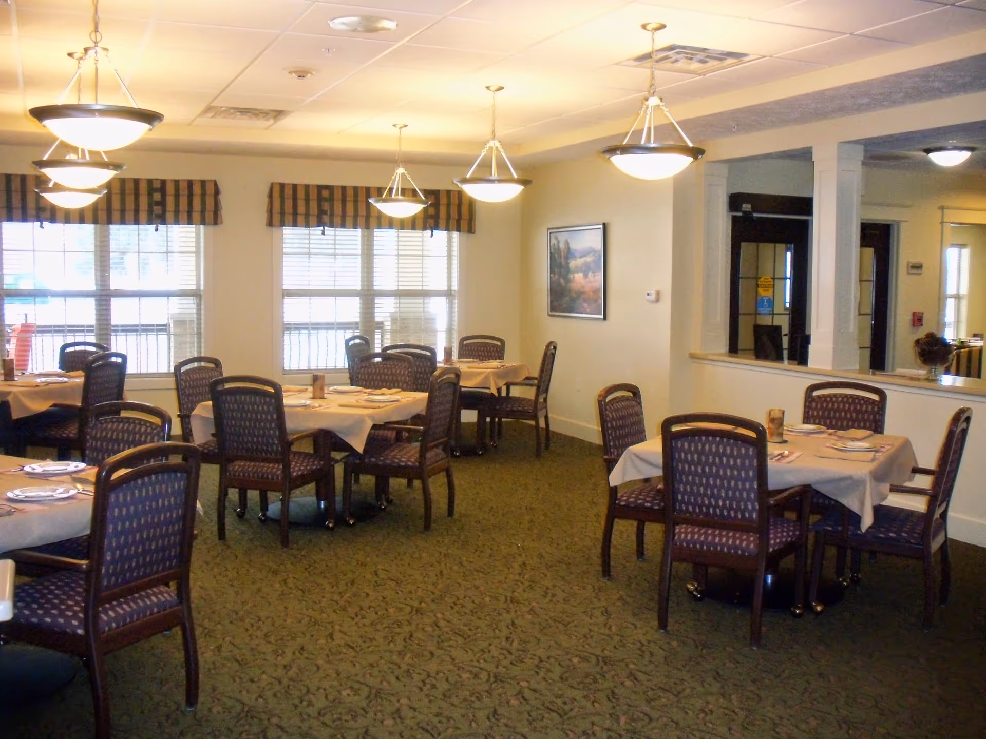Dining room with multiple tables set for meals, chairs, windows, and pendant lights.