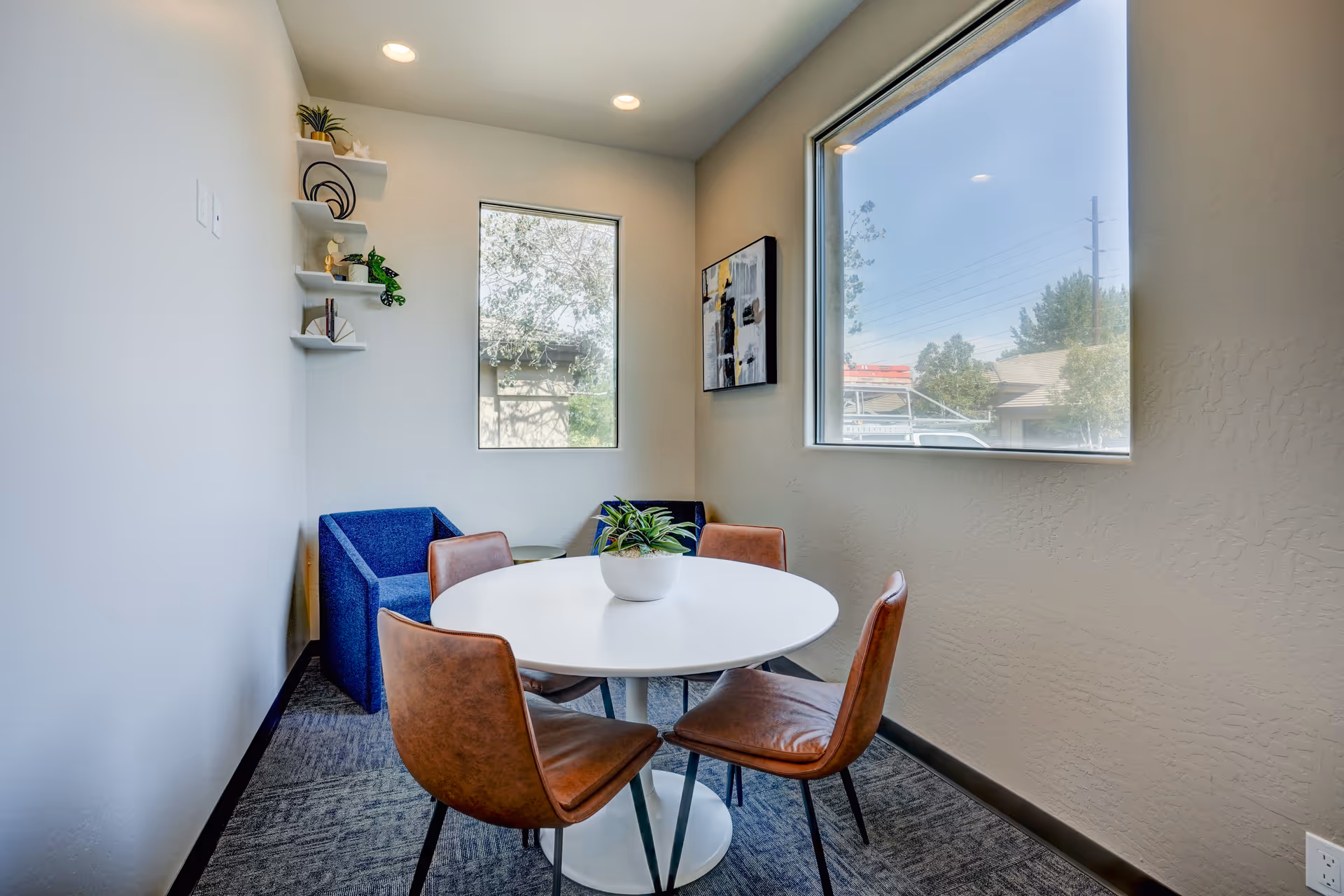 Small bright interior lounge with round table, four chairs, and wall shelves by a large window.
