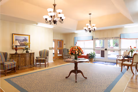 A bright and spacious living room area with a large patterned rug, a round wooden table with a vase of flowers in the center, several upholstered chairs, a wooden sideboard with a lamp and framed artwork above it, and large windows and glass doors letting in natural light.