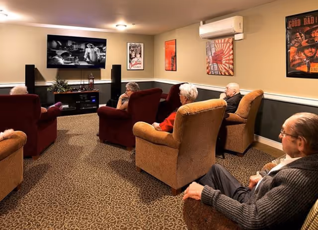 Several elderly residents seated in armchairs watching a movie on a wall-mounted TV in a cozy common lounge.