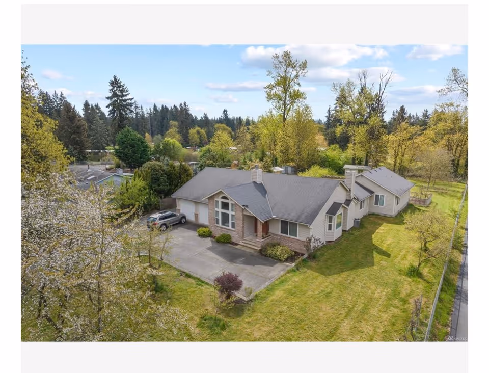 Aerial view of a single-story residential building surrounded by green lawns and trees under a partly cloudy sky. The house has a gray roof, beige siding, and a driveway with a parked car. The property is fenced and located in a wooded area.