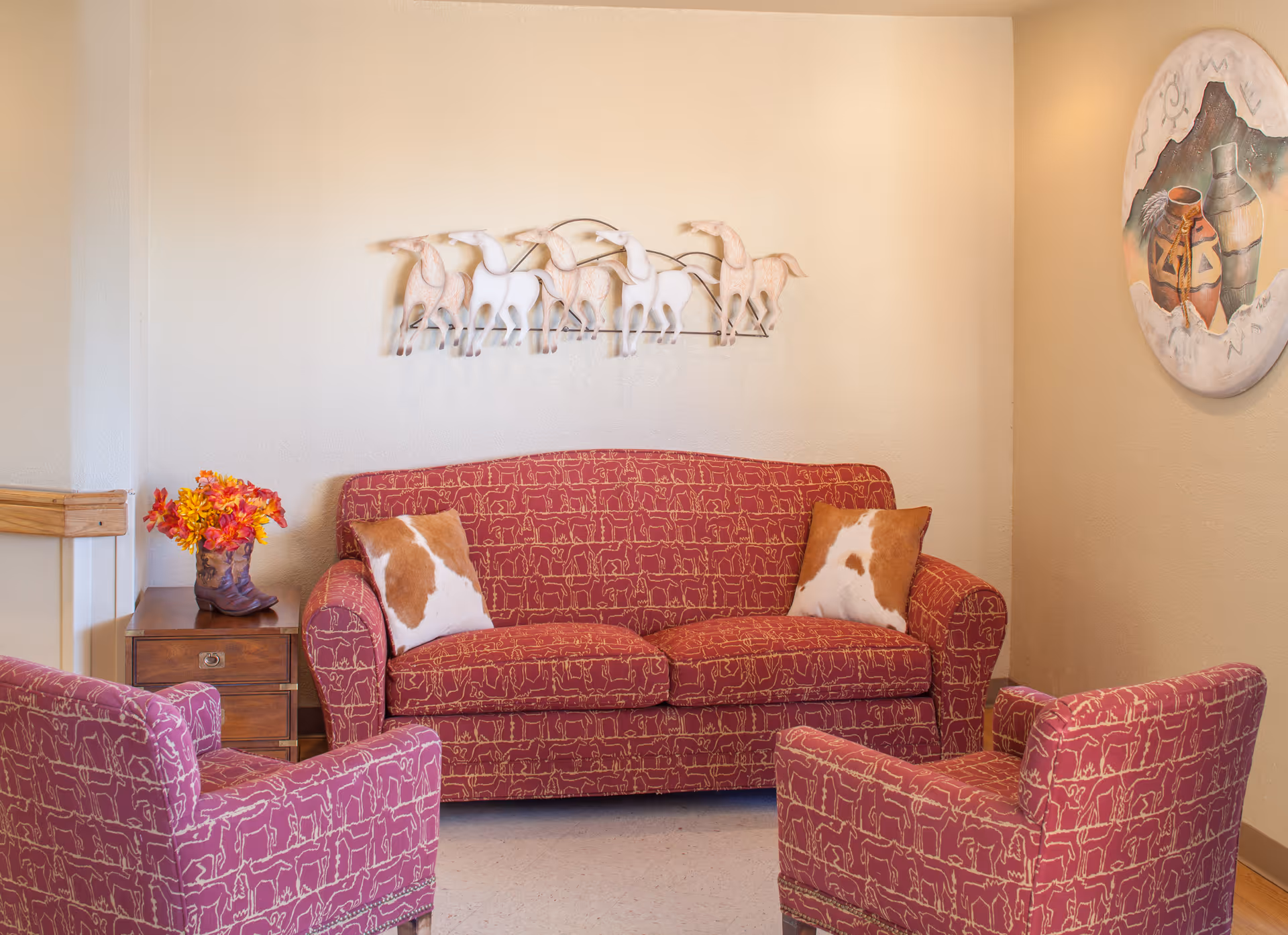 A cozy seating area with a patterned red sofa and two matching armchairs, cowhide pillows, wall art, and a small side table with flowers.