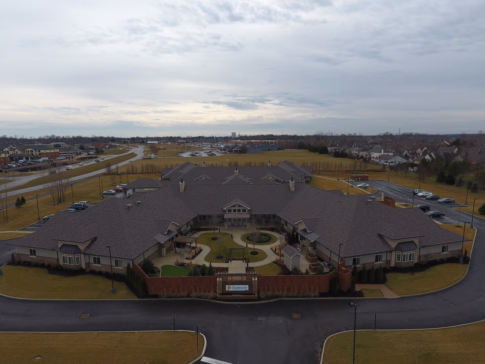 Aerial view of The Ganzhorn Suites senior living facility showing a large, single-story building with a central courtyard, surrounded by parking lots and open grassy areas under a cloudy sky.