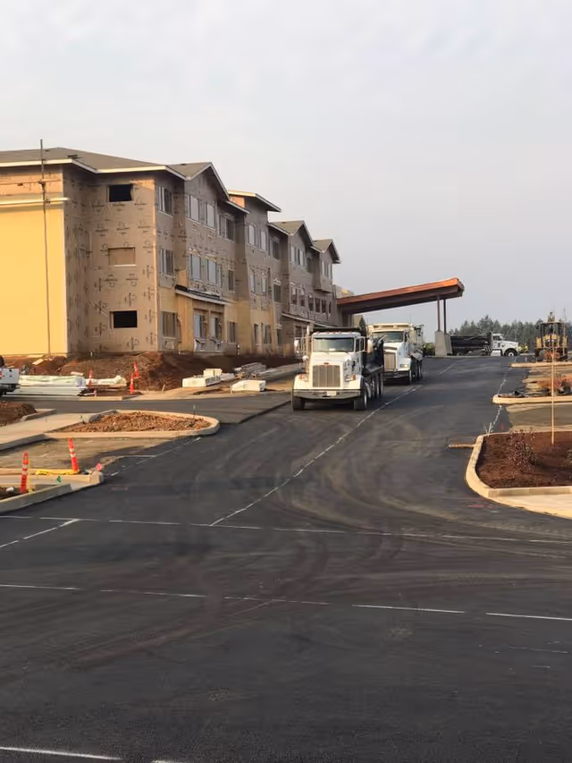 A large multi-story building under construction with trucks parked on a newly paved road in front. The building is partially covered with construction wrap and has a covered entrance. The surrounding area shows construction materials and landscaping work in progress.