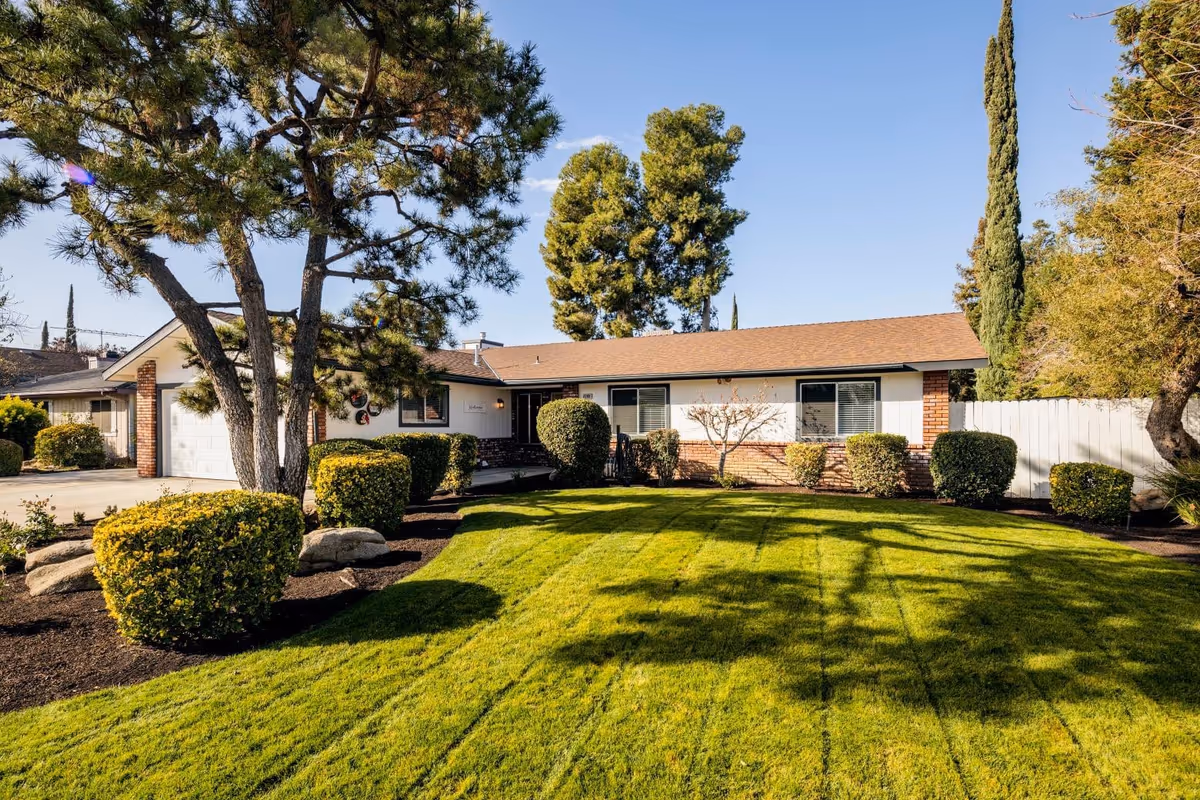Single-story house with manicured lawn, trimmed shrubs, and large trees in front yard.
