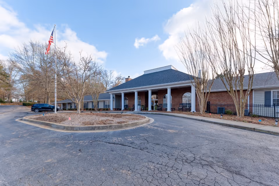 Exterior view of Addington Place of Roswell, a single-story brick building with white columns at the entrance, a circular driveway, and an American flag on a flagpole. Leafless trees and a parked black SUV are visible under a partly cloudy sky.