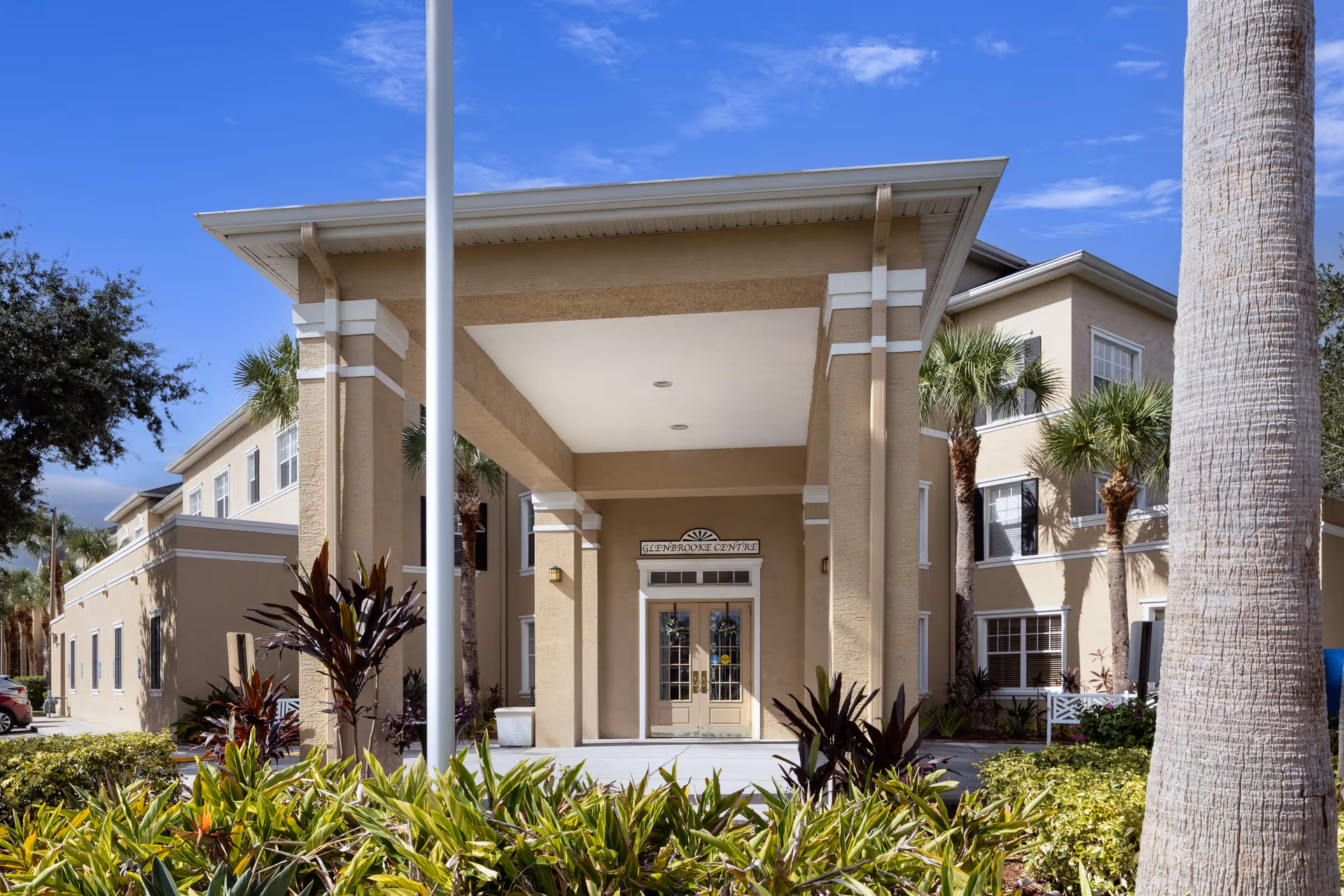 Exterior front entrance of Glenbrooke Senior Living facility with beige walls, large covered entryway, glass double doors, palm trees, and landscaped greenery under a blue sky.
