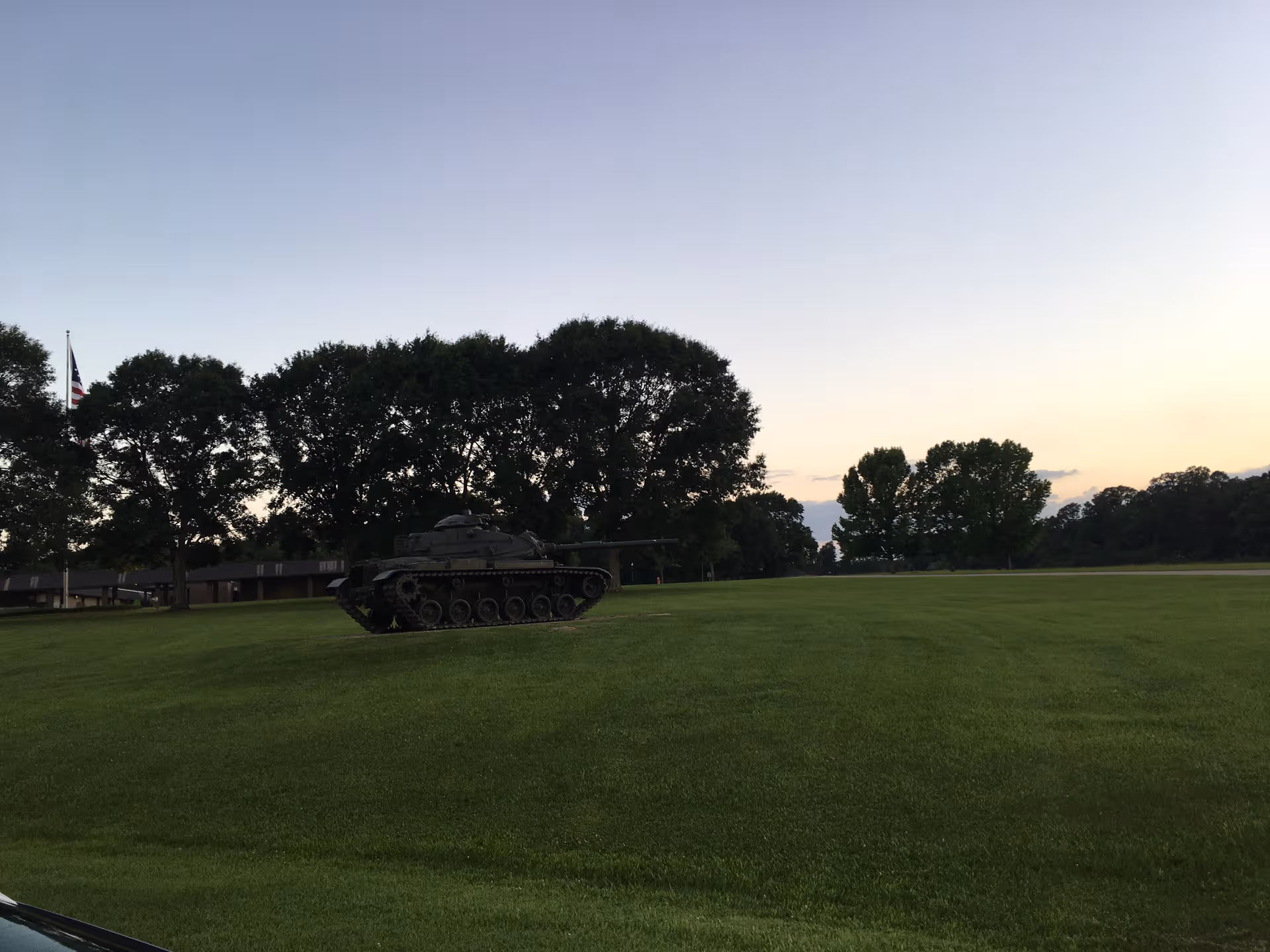 An outdoor lawn with a military tank monument, trees, and a flag near a low building.