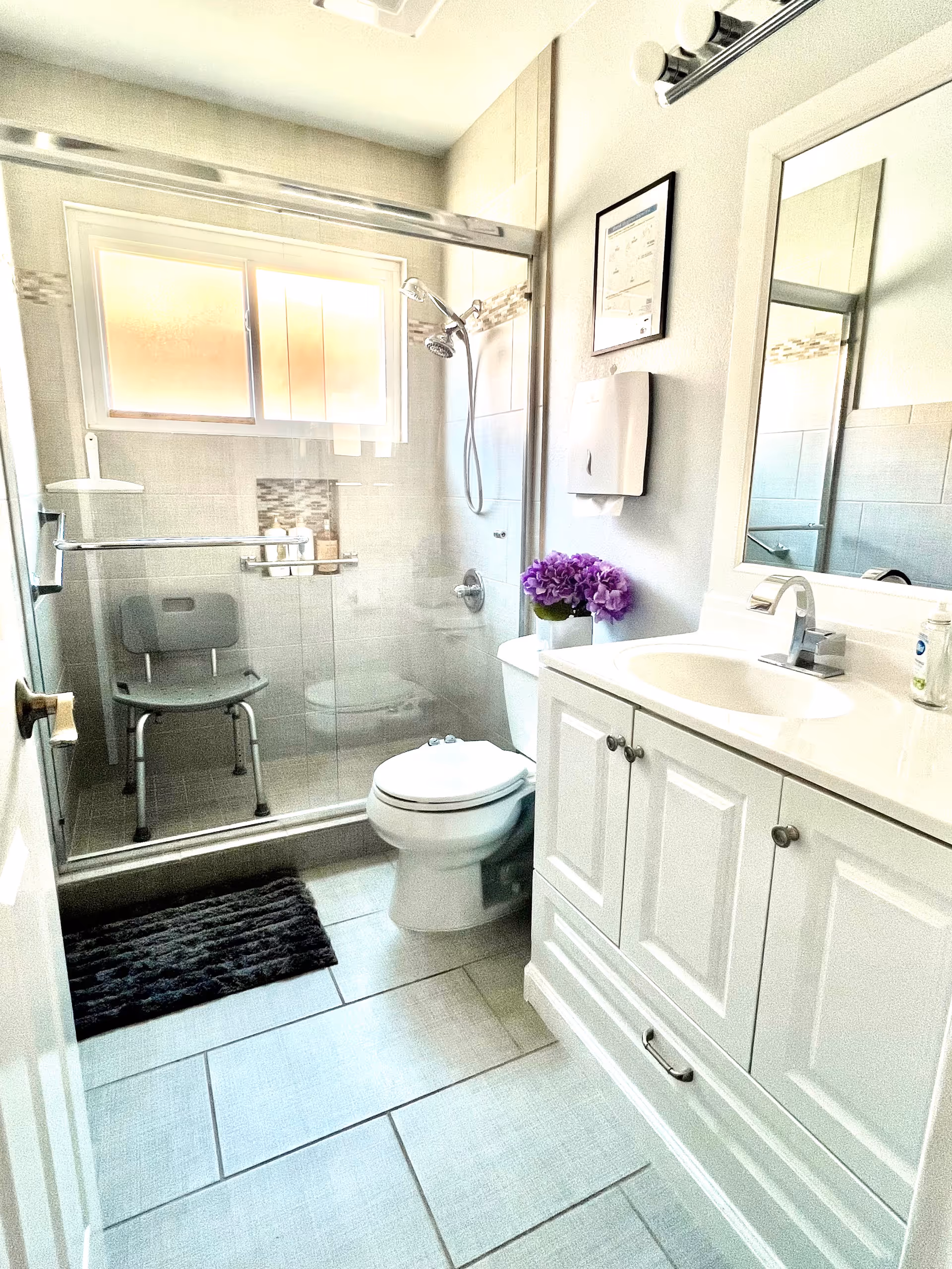Bright bathroom with a glass shower, toilet, white vanity, and purple flowers.