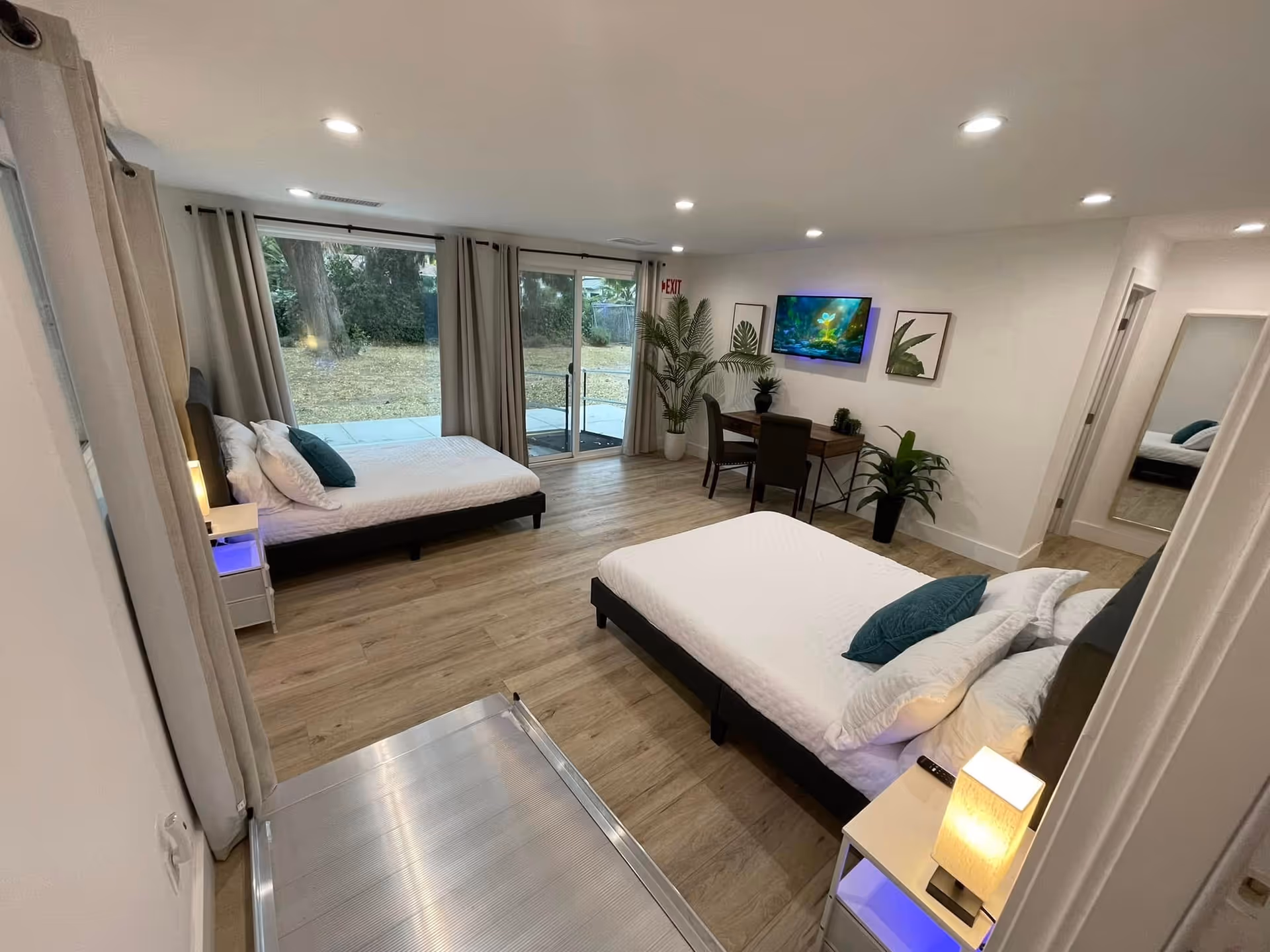 Bright senior living bedroom suite with two beds, TV, desk, plants, and sliding glass door.