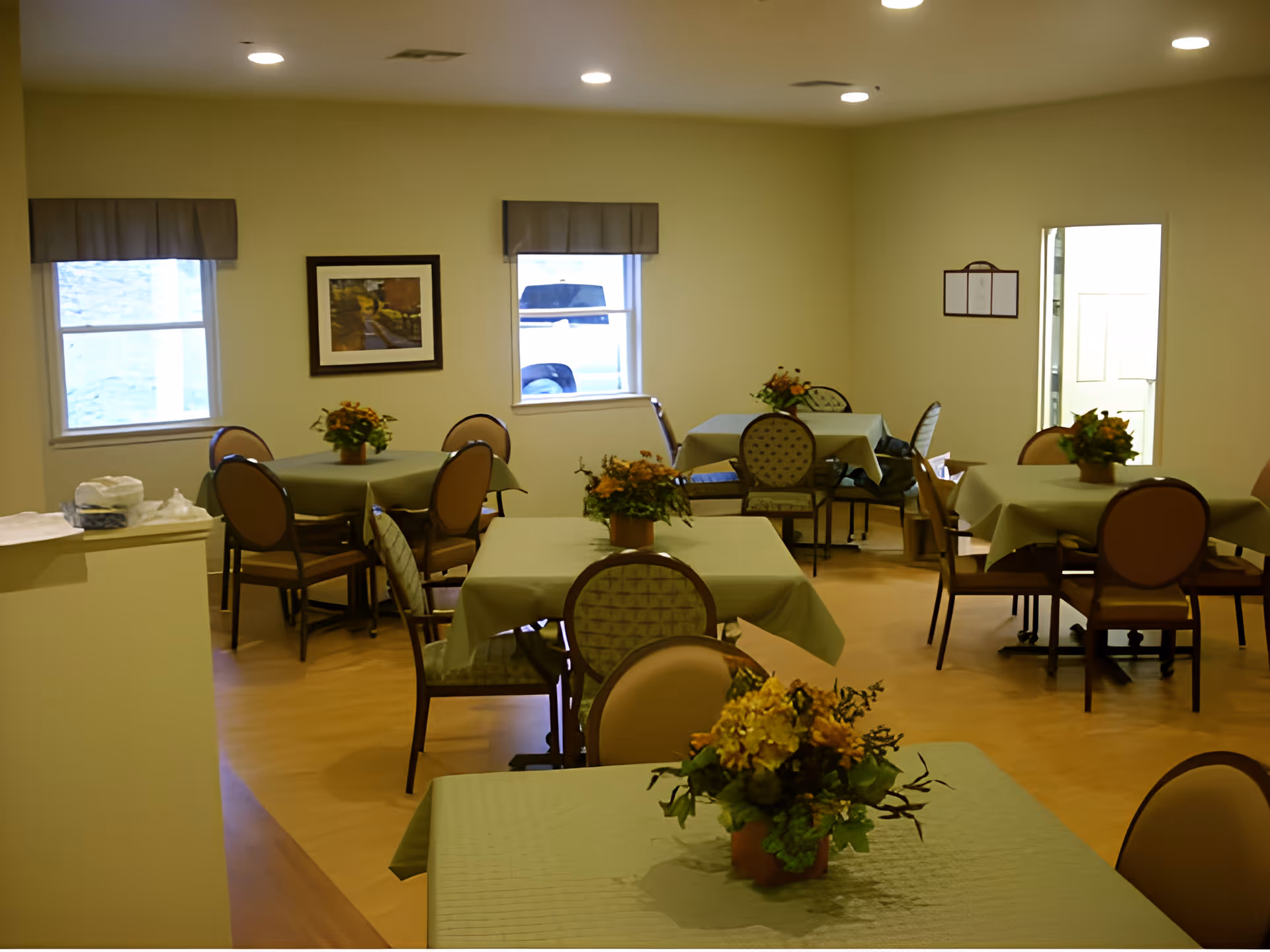 A dining room with several tables covered in green tablecloths, each adorned with a floral centerpiece. The room has beige walls, two windows with valances, a framed picture on the wall, and several chairs around the tables. The floor is wooden, and there is a doorway visible on the right side.