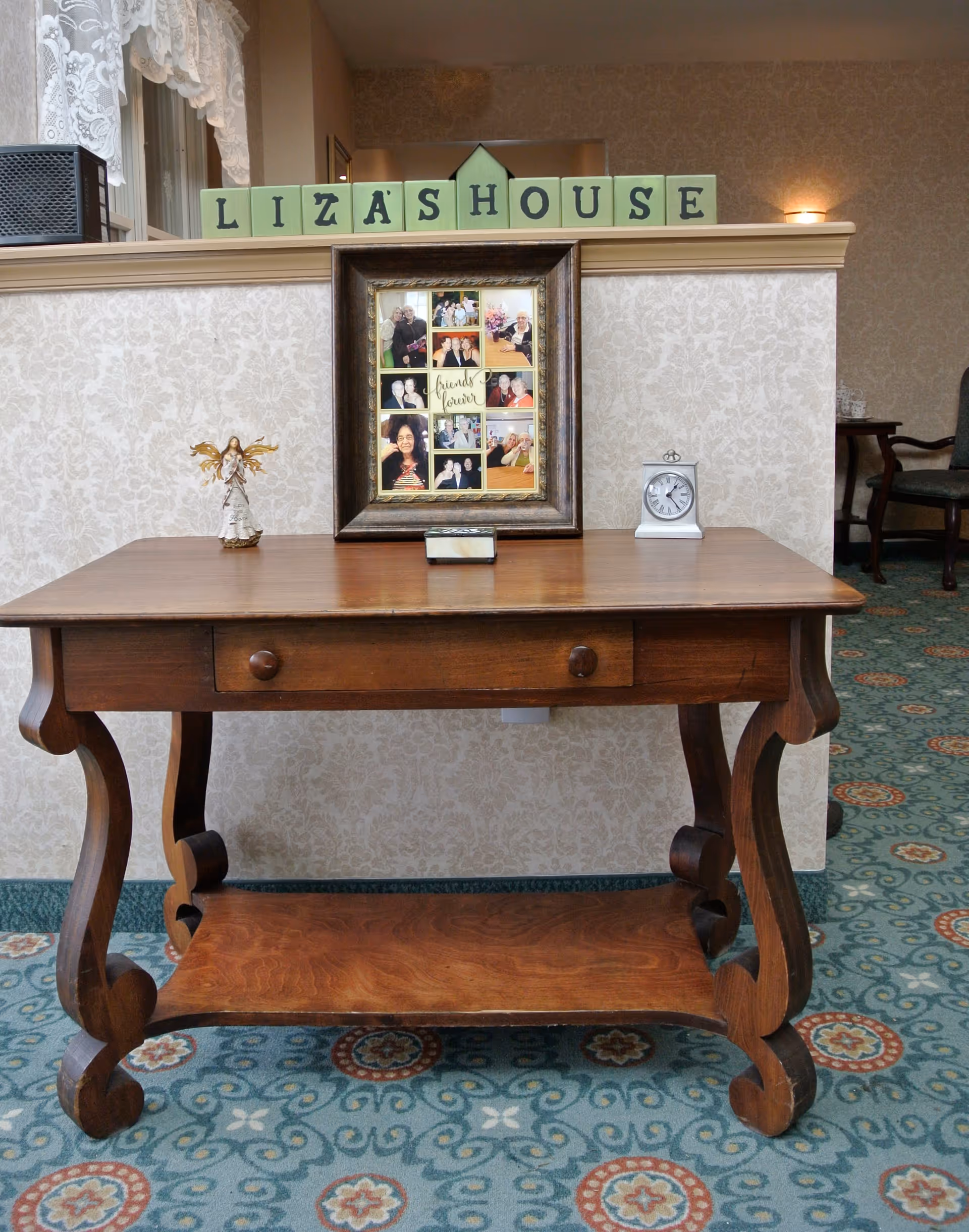 A wooden table with curved legs and a drawer, placed against a wallpapered half wall. On the table, there is a small angel figurine, a framed collage of photos, and a small white clock. Above the wall, green blocks spell out 'LIZA'S HOUSE'. The floor is covered with a patterned carpet in blue and red tones.