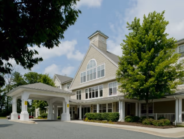 Exterior front entrance of a multi-story senior living facility with a covered porte-cochere, large windows, and landscaped trees.