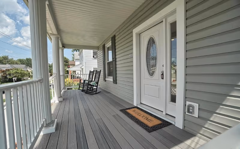 Covered front porch with rocking chairs, white door, welcome mat, grey siding.