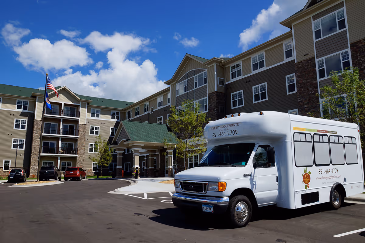 Exterior front of a multi-story senior living building with a parked white shuttle bus near the entrance under a blue sky.